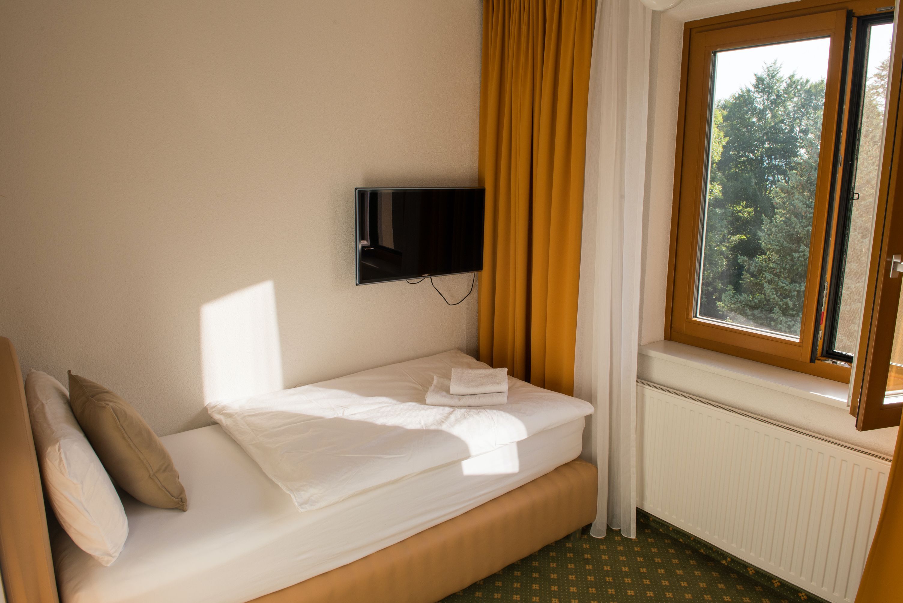 Einzelzimmer mit Einzelbett, gelben Vorhängen, einem Fenster mit Blick ins Grüne und einem an der Wand montierten Fernseher.