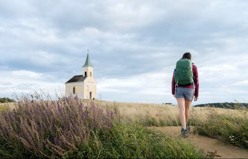 Wanderer durchqueren die sanften Hügel, umgeben von hohen Gräsern, die im Wind wiegen. Die kleine Kapelle auf dem Hügel strahlt Ruhe und Spiritualität aus und lädt dazu ein, einen Moment innezuhalten und die atemberaubende Aussicht zu genießen.
