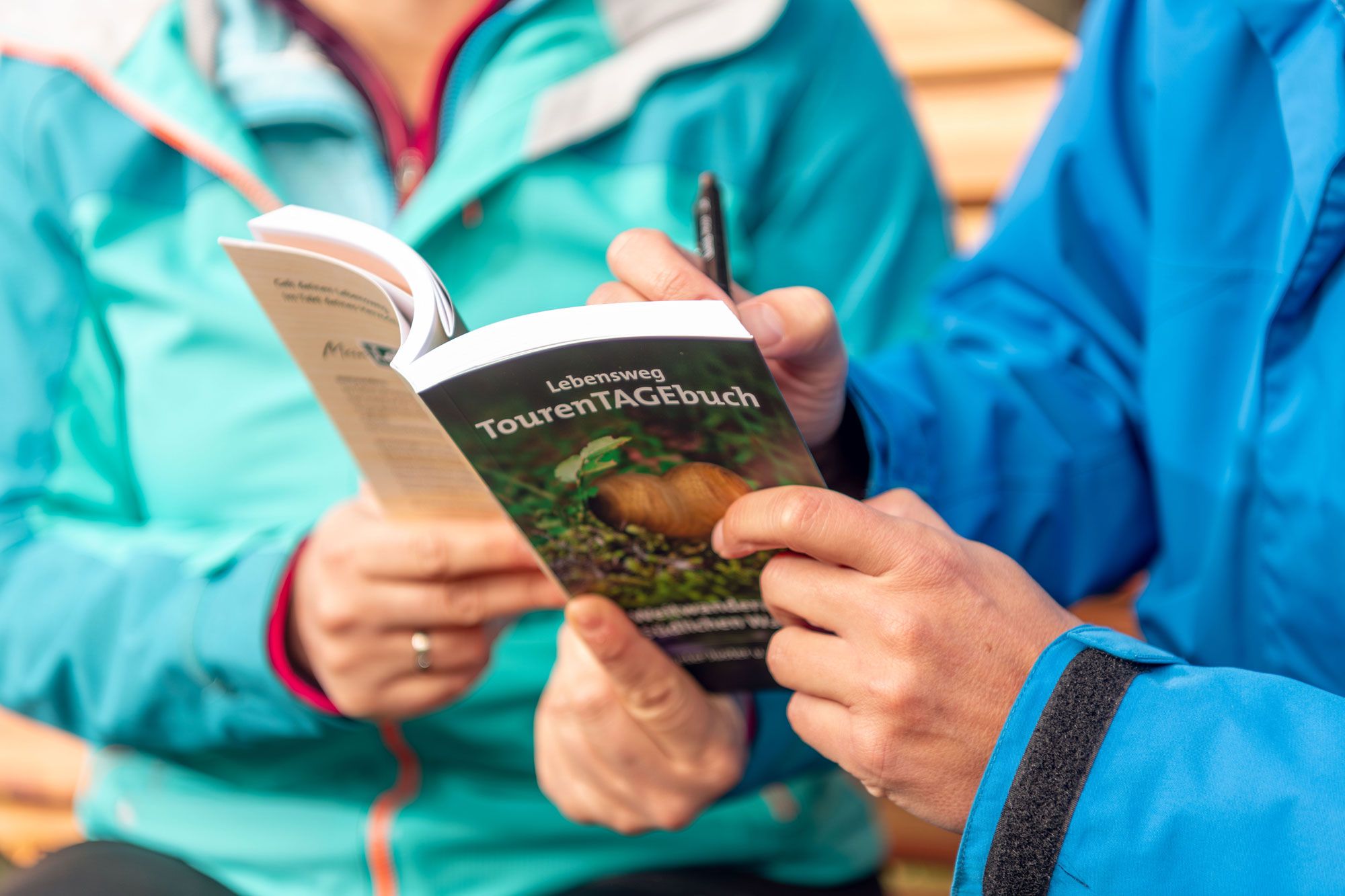 Zwei Personen in blauen Jacken halten ein Buch mit dem Titel 'Lebensweg TourenTAGEbuch'.