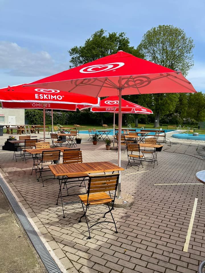 Ein Freibad mit roten Sonnenschirmen und leeren Holztischen auf einer gepflasterten Terrasse.