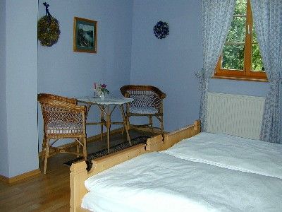 Ein gemütliches Zimmer mit zwei Betten, einem kleinen Tisch und zwei Stühlen. Fenster mit Vorhängen und Wanddekorationen sind sichtbar.