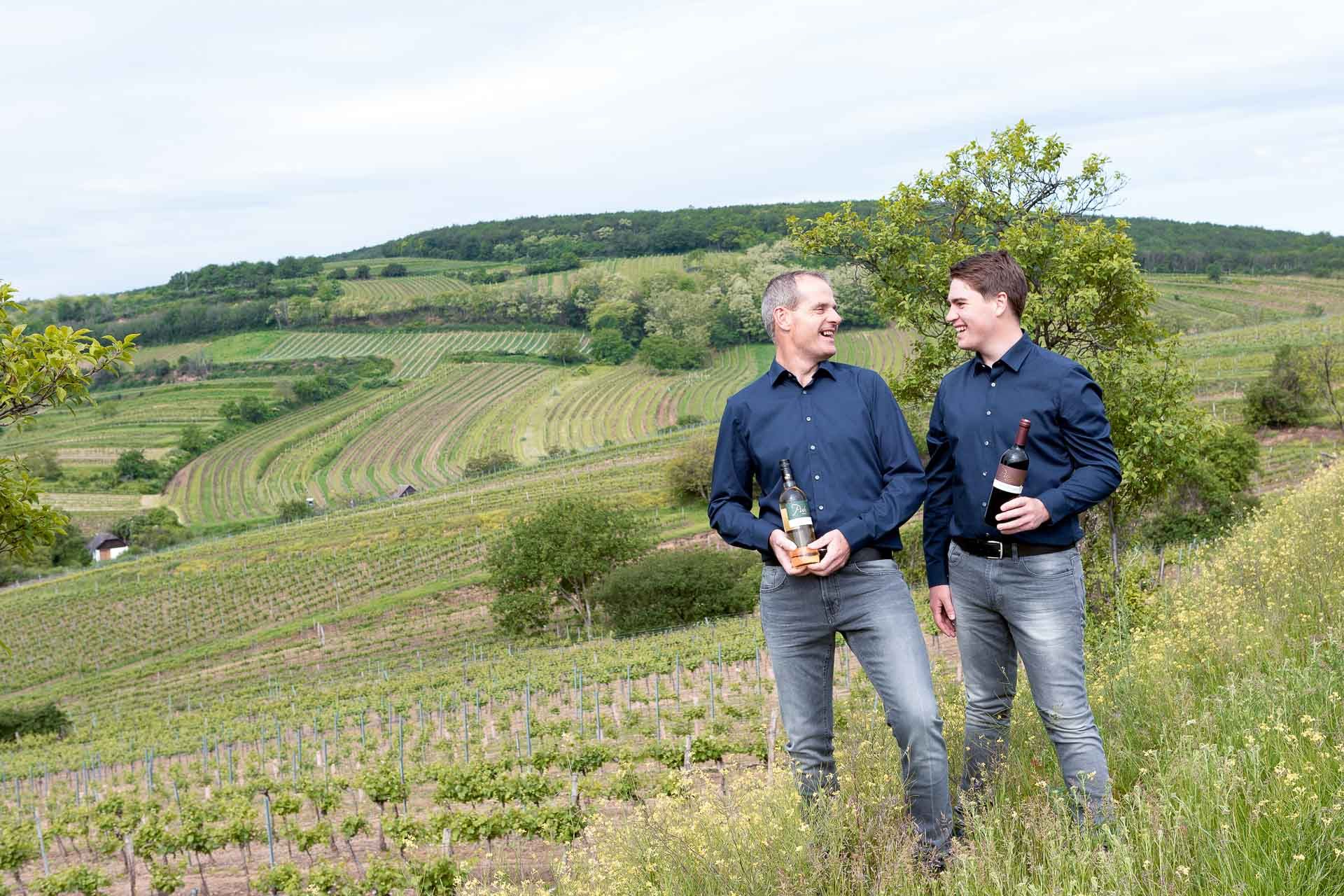 Zwei Männer in blauen Hemden stehen in einem Weinberg und halten Weinflaschen.