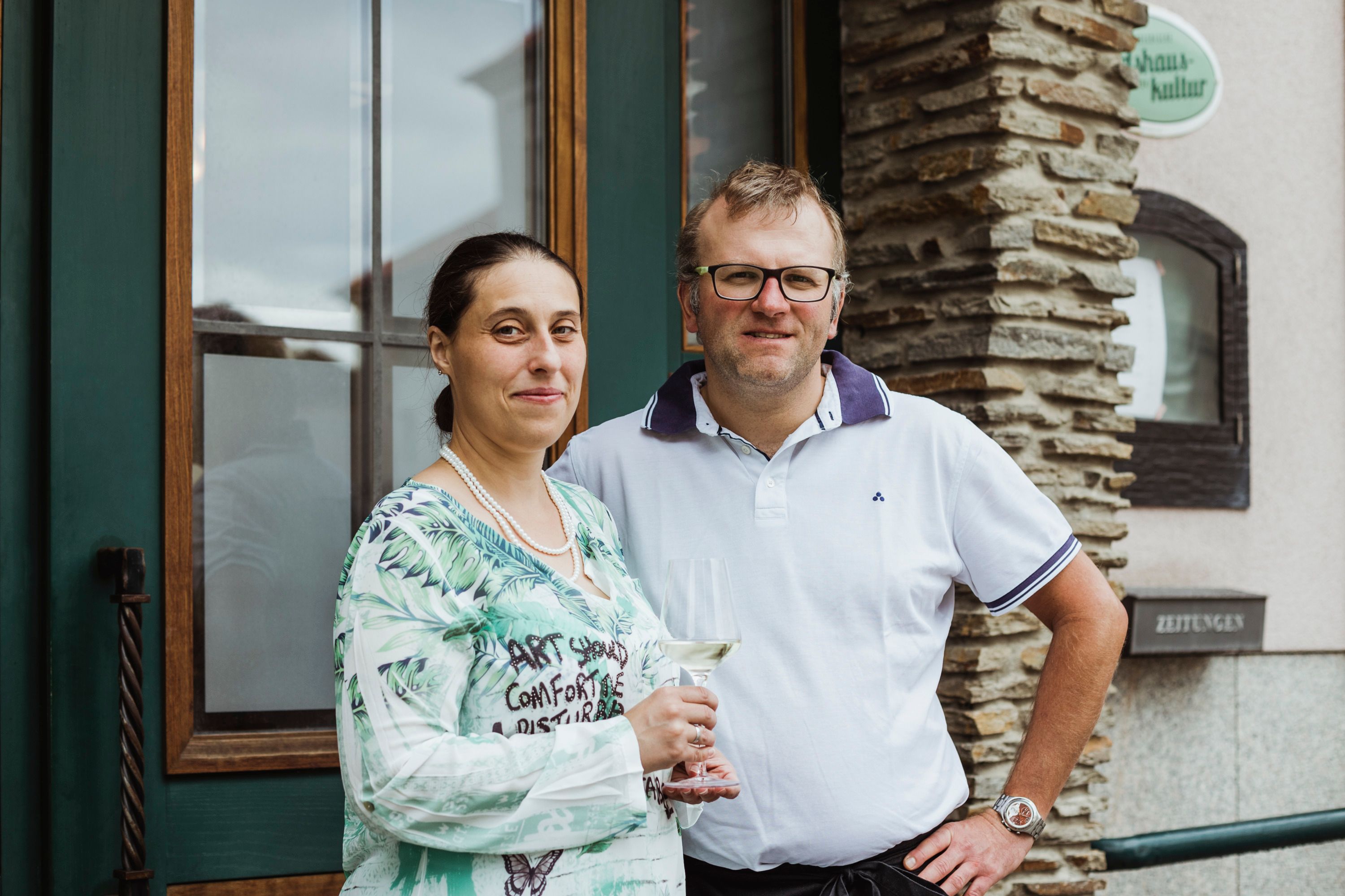 Ein Mann und eine Frau stehen lächelnd vor einem Gebäude. Die Frau hält ein Weinglas in der Hand.
