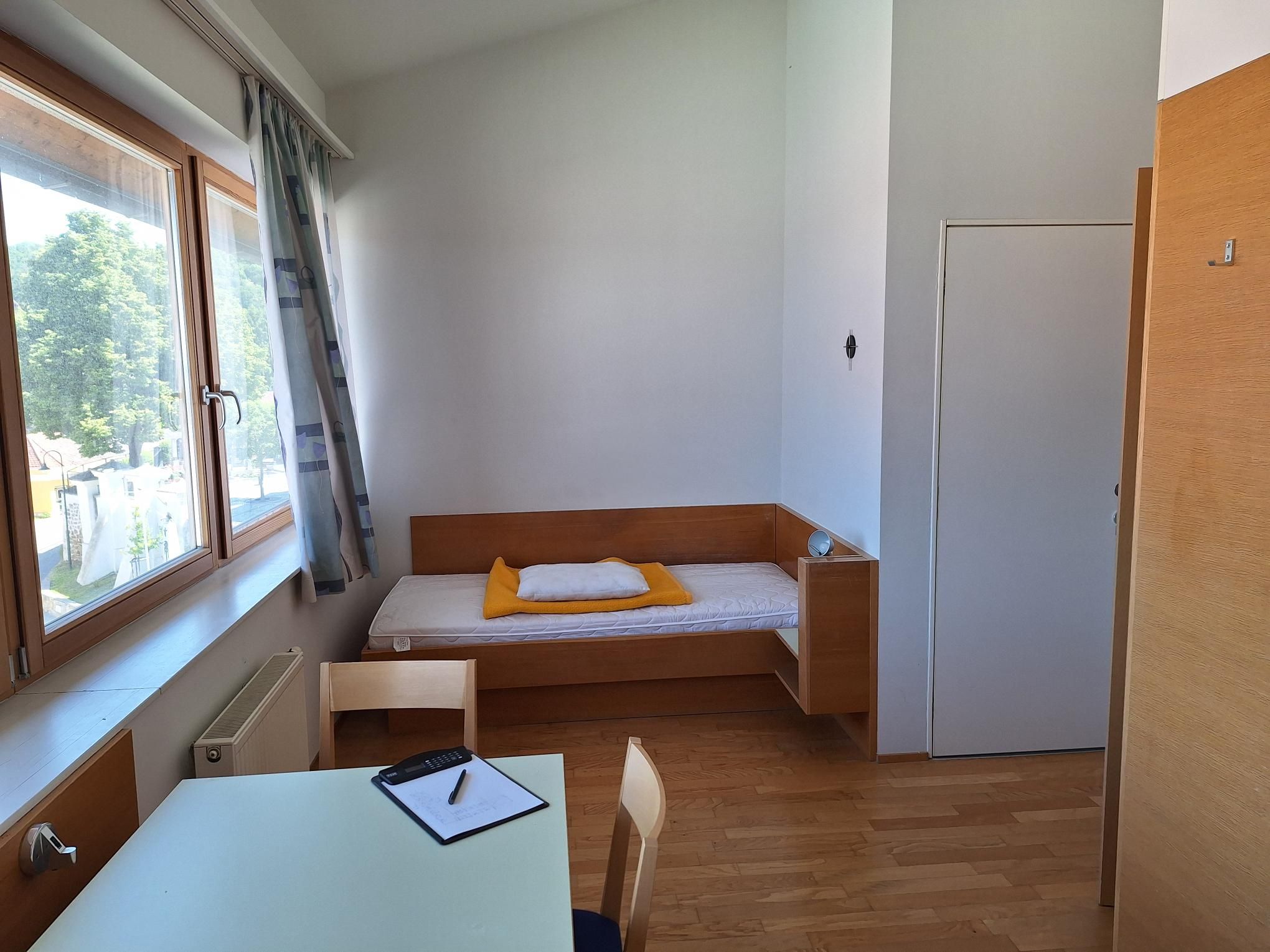 Ein einfach eingerichtetes Einzelzimmer mit Bett, Tisch und Stuhl, helle Holzmöbel und ein großes Fenster mit Blick nach draußen.