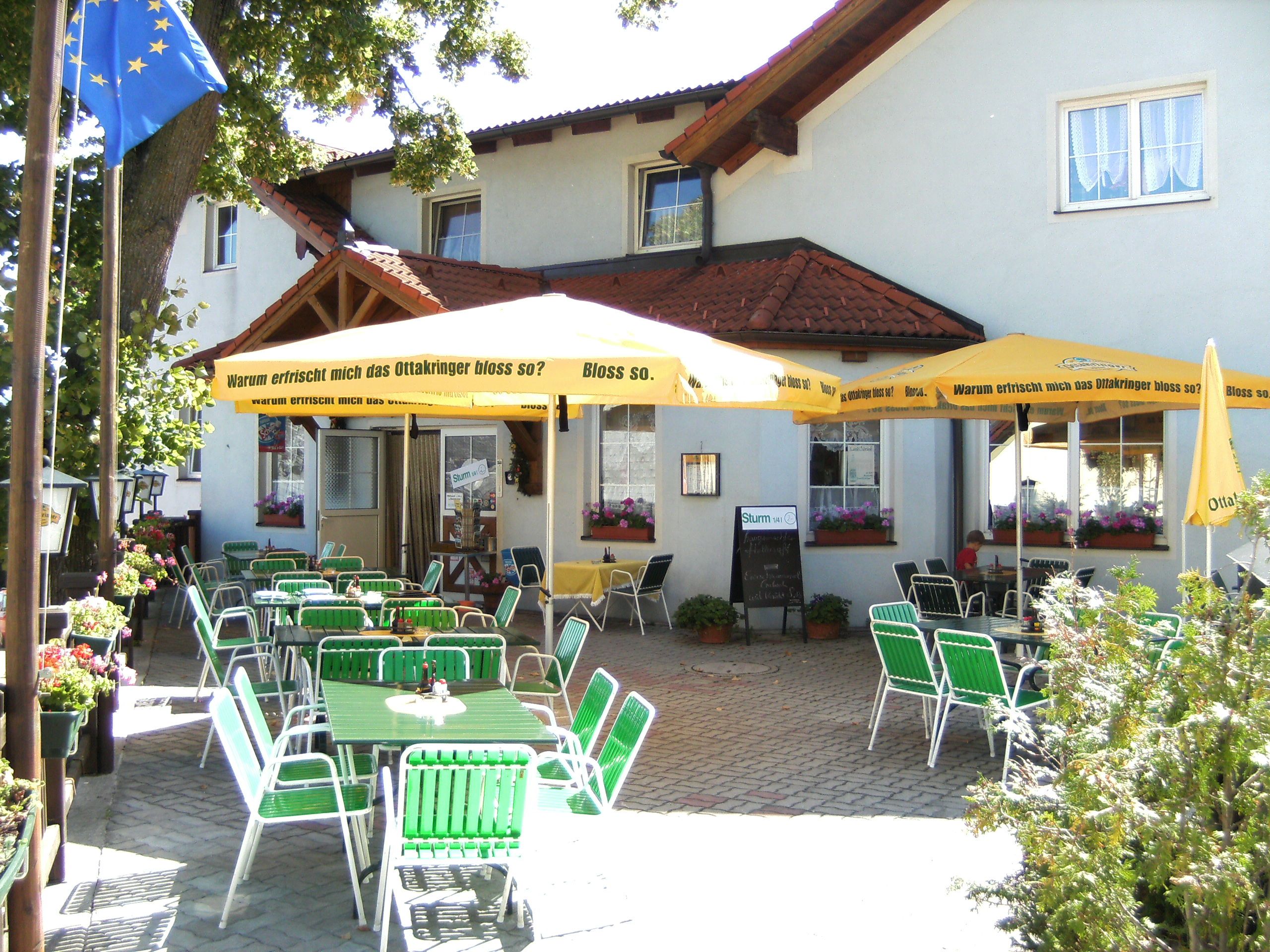 Ein Gastgarten mit grünen Stühlen und gelben Sonnenschirmen vor einem Gebäude.