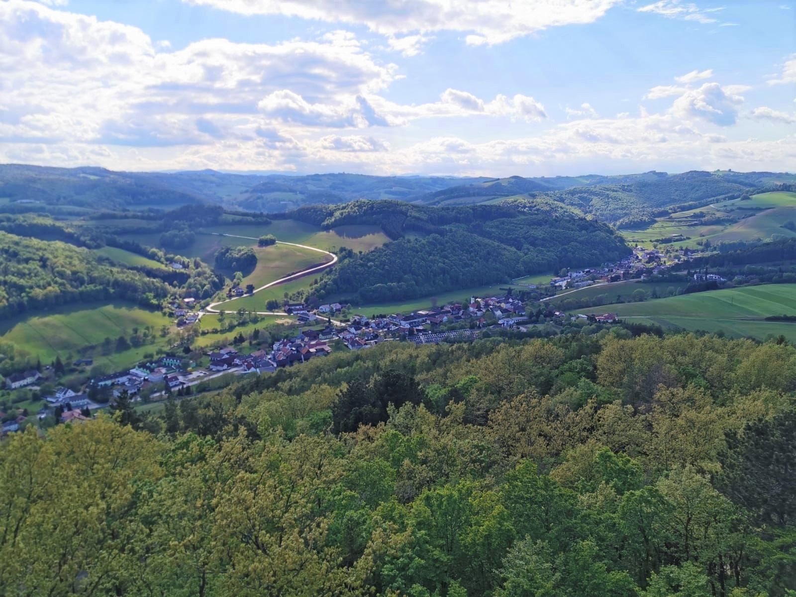 Panoramablick auf grüne Hügellandschaft mit Wäldern, blauem Himmel und kleinem Dorf.
