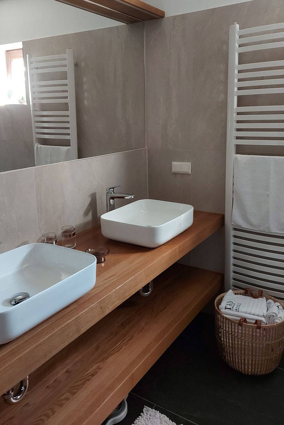 Modernes Badezimmer mit zwei weißen Waschbecken auf Holzplatte, Handtuchhalter und Korb mit Handtüchern.