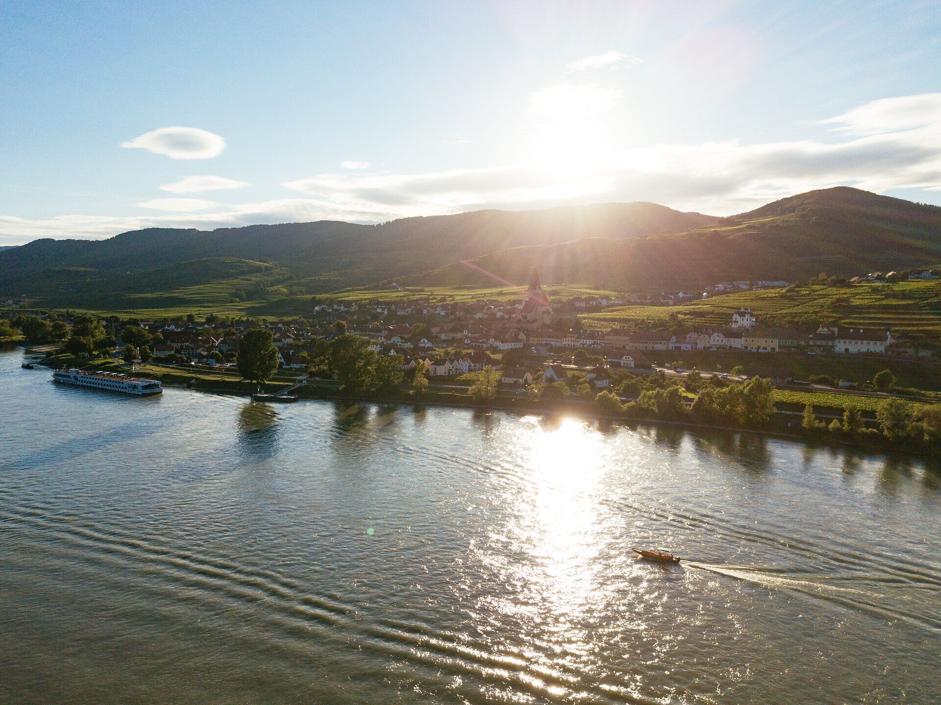 Die sanften Hügel der Wachau erstrahlen im warmen Licht der Abendsonne, während die Donau friedlich dahinfließt. Ein Boot gleitet über das glitzernde Wasser und lädt dazu ein, die atemberaubende Landschaft zu genießen, die von Weinbergen und charmanten Dörfern geprägt ist.