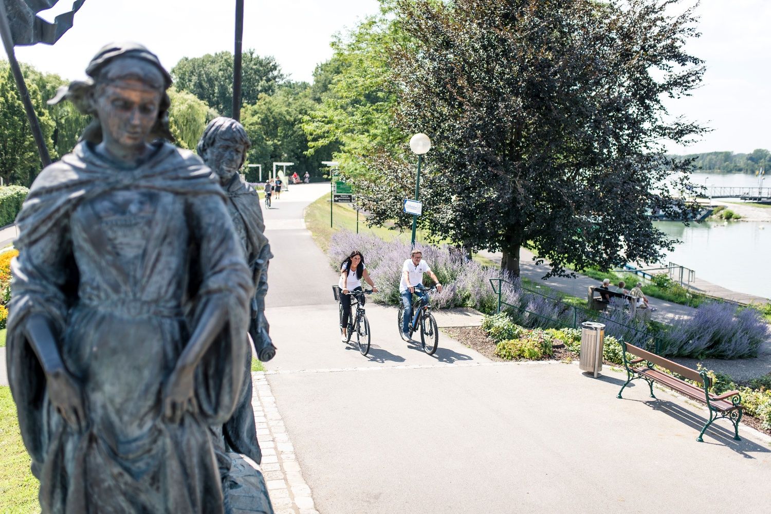 Zwei Personen fahren auf einem Radweg an einer Statue vorbei, umgeben von Bäumen und einem Fluss im Hintergrund.