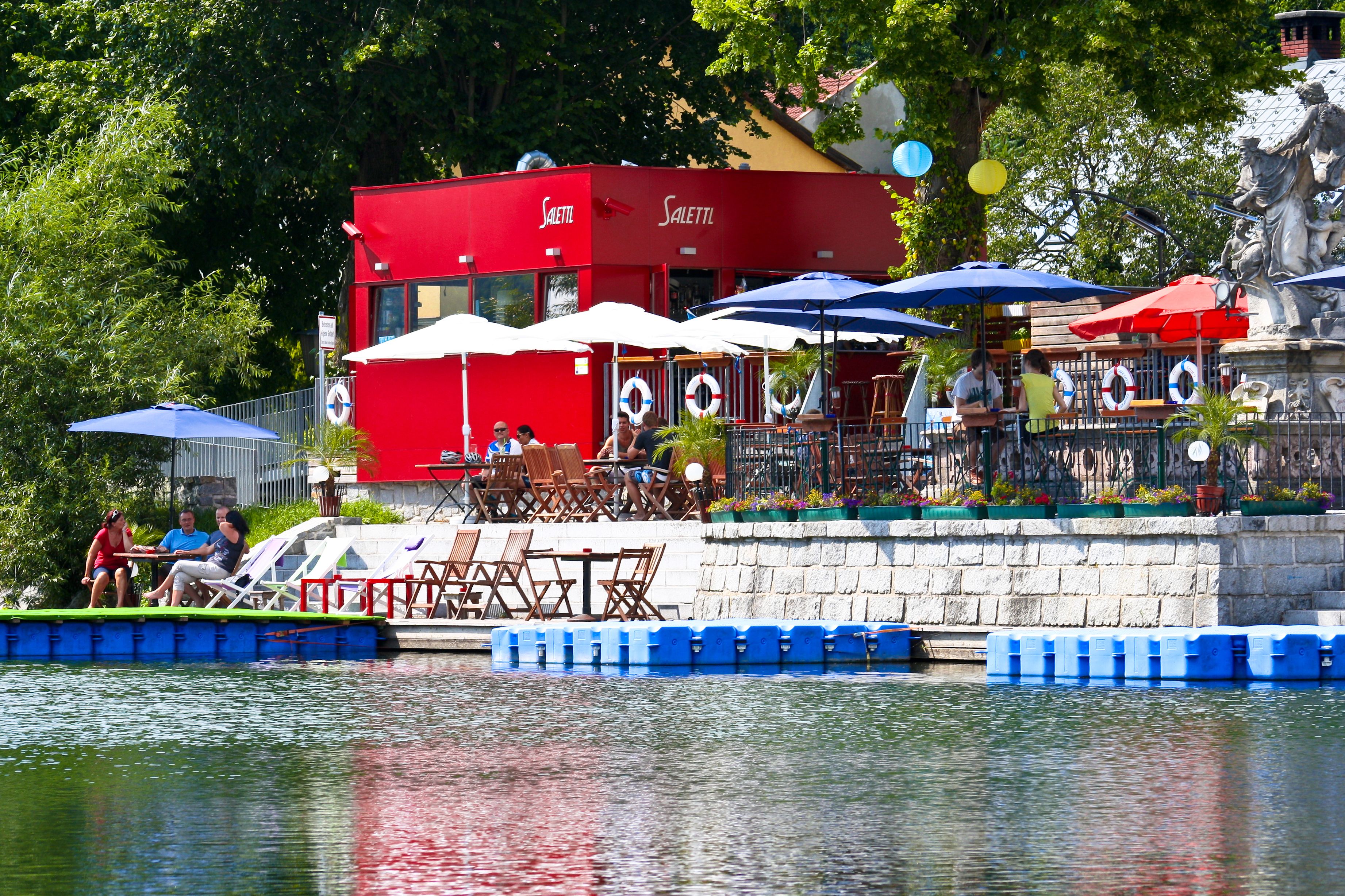Ein rotes Gebäude mit Terrasse am Wasser, umgeben von Sonnenschirmen und Sitzgelegenheiten.