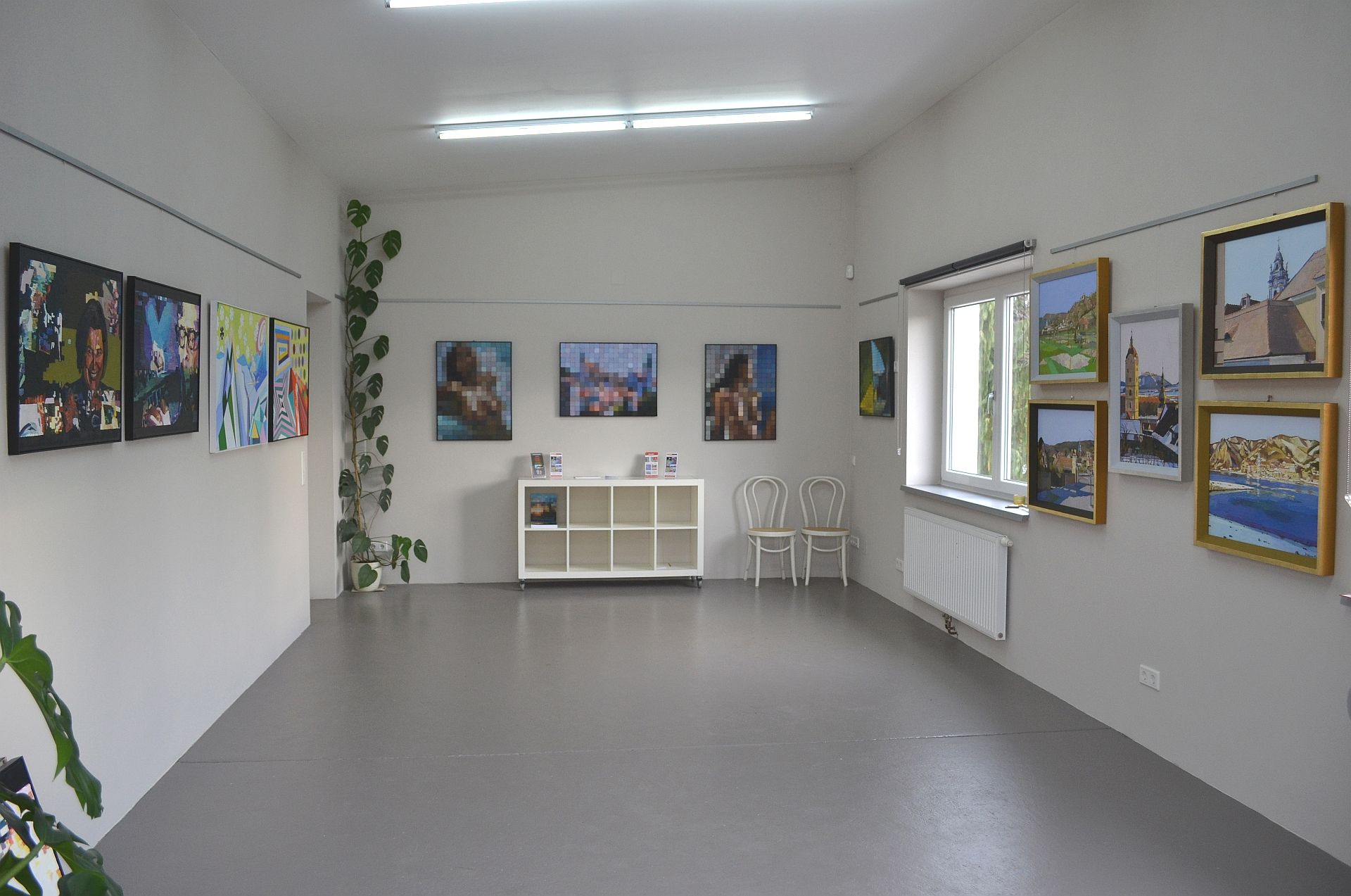 Innenansicht einer Galerie mit Gemälden an den Wänden und einem Regal in der Mitte.