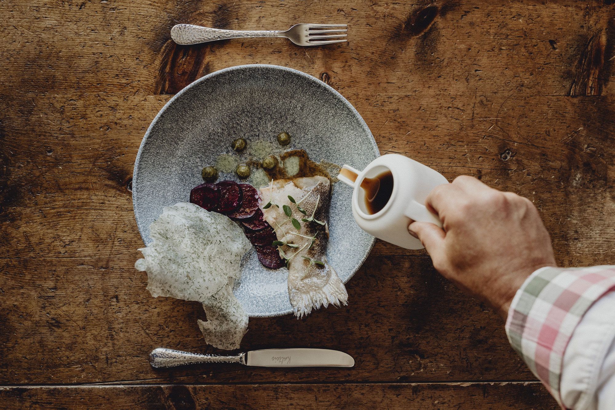 Ein Teller mit Fisch, Roter Bete und Kapern auf einem Holztisch. Eine Hand gießt Sauce aus einer Kanne darüber.