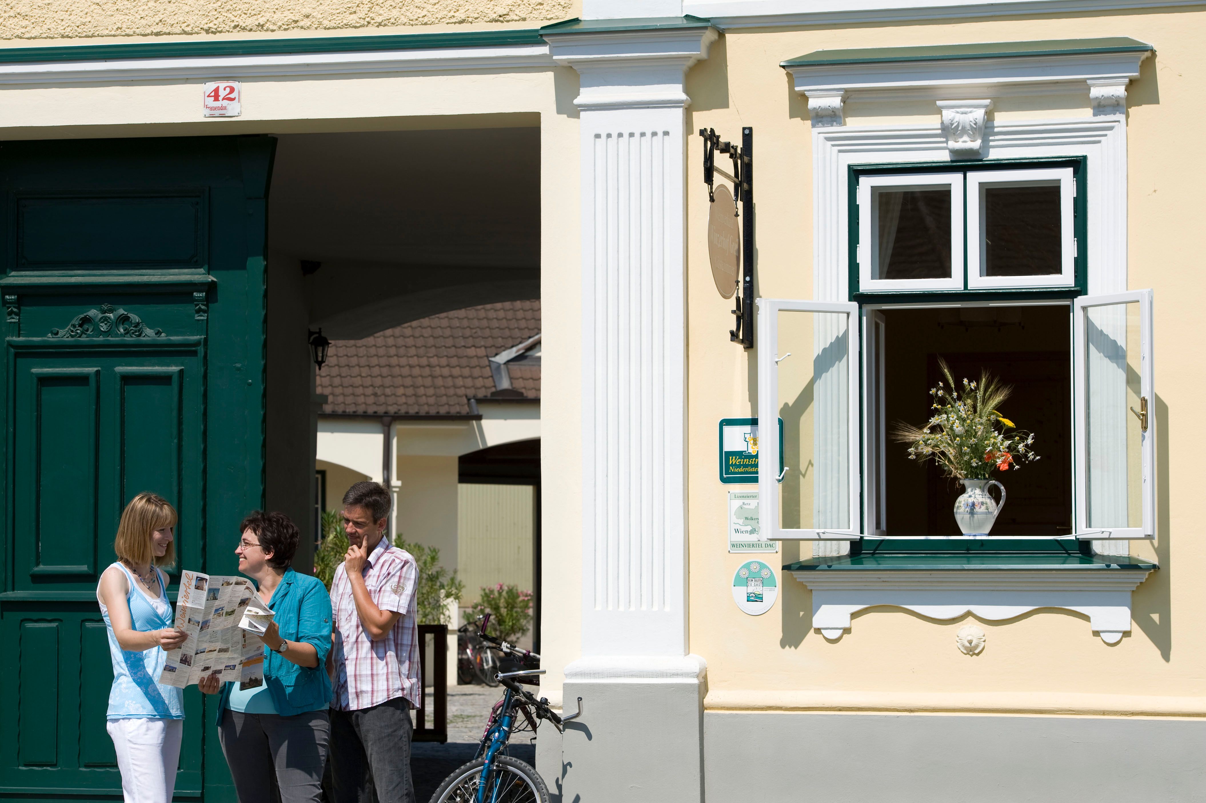 Drei Personen stehen vor einem gelben Gebäude mit grüner Tür und lesen eine Karte. Ein Fenster mit Blumenstrauß ist geöffnet.