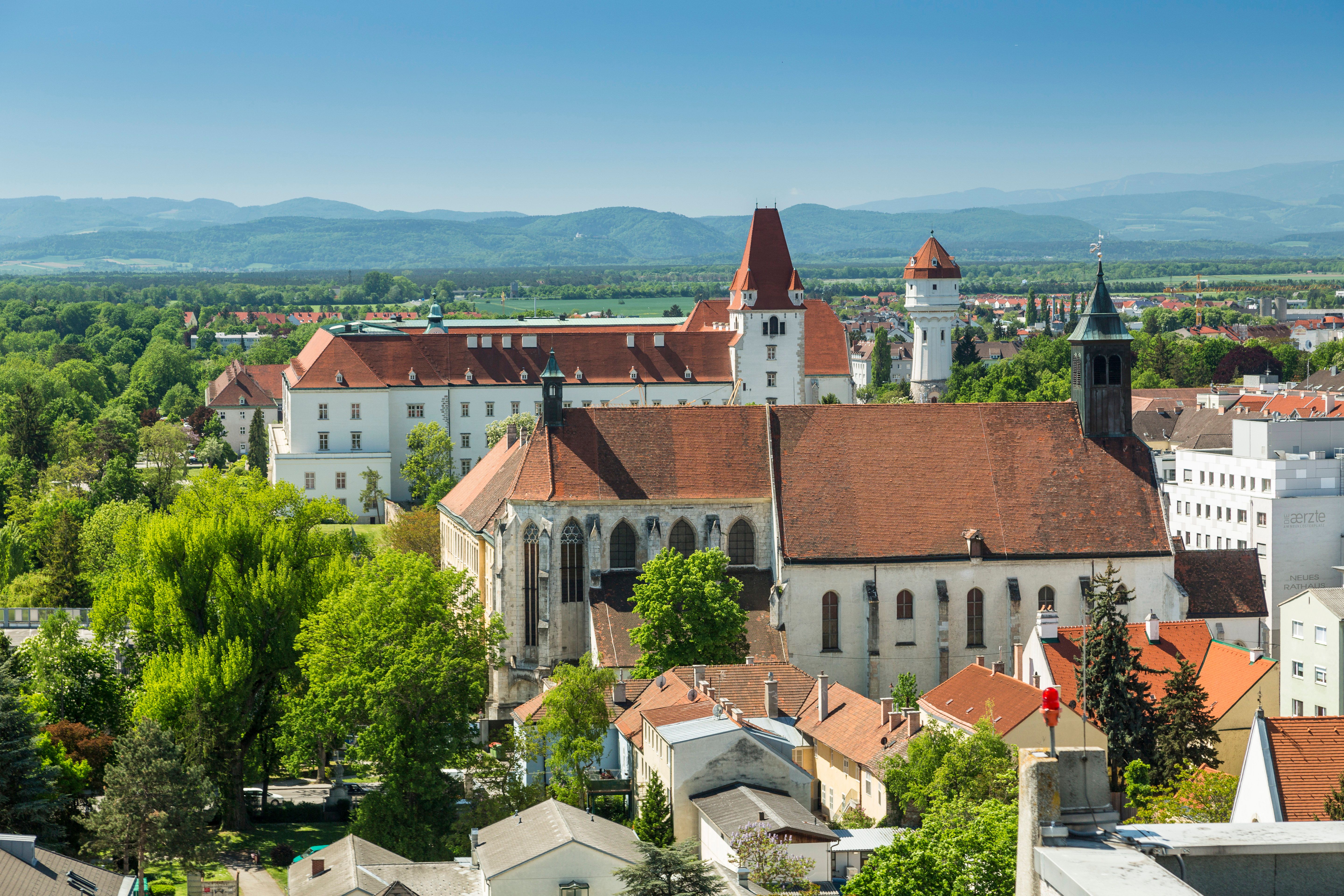 Drei Sehenswürdigkeiten auf einem Foto: Das Stift Neukloster, die Theresianische Militärakademie am Akademiepark und der Wasserturm. Im Hintergrund die Berge der Wiener Alpen.
