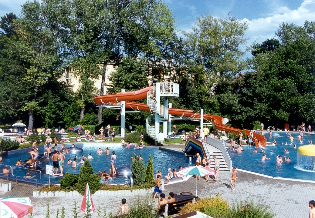 Ein Freibad mit vielen Menschen, einer Wasserrutsche und Bäumen im Hintergrund.