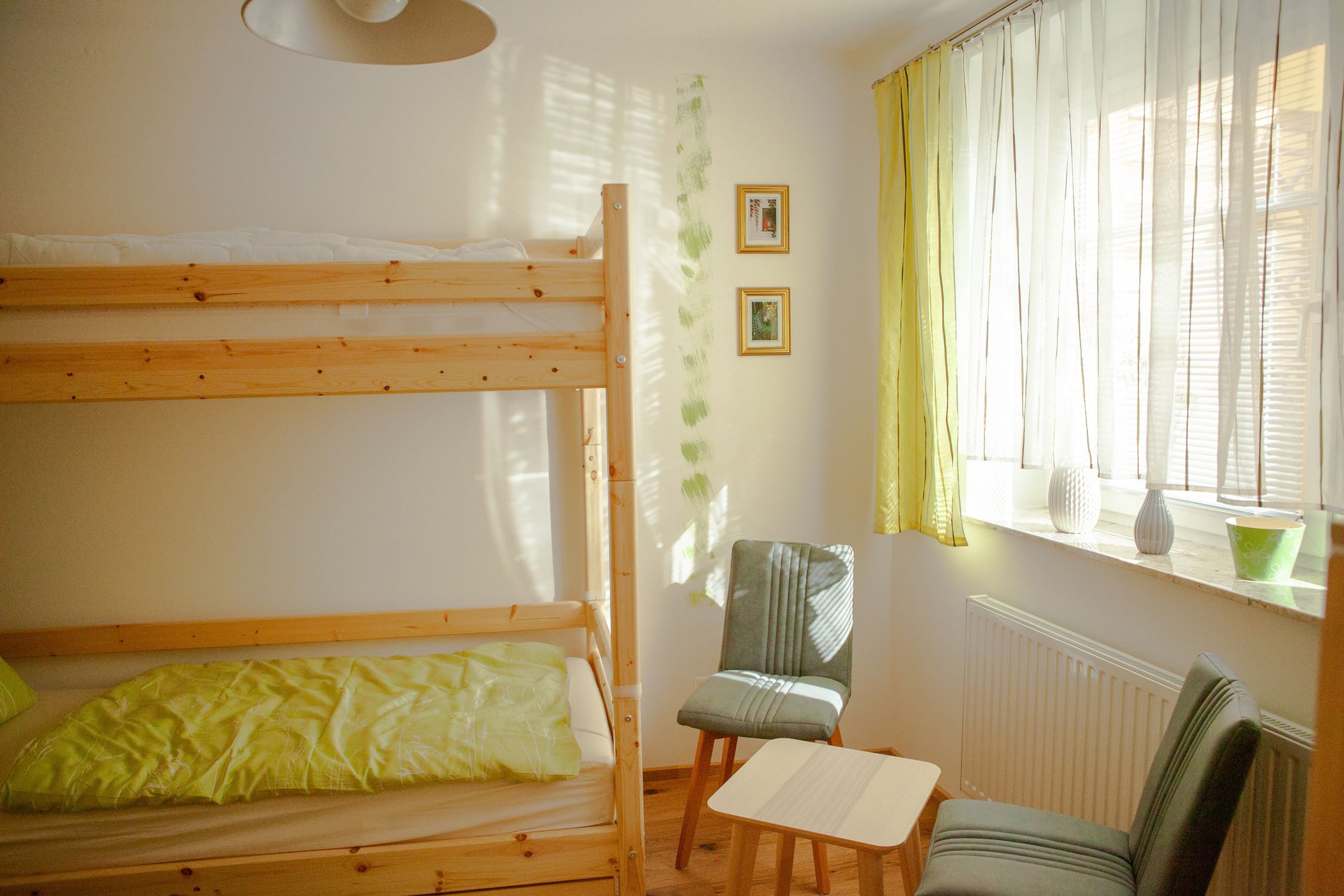 Ein helles Zimmer mit einem Stockbett, zwei Stühlen, einem kleinen Tisch und einem Fenster mit Vorhängen.
