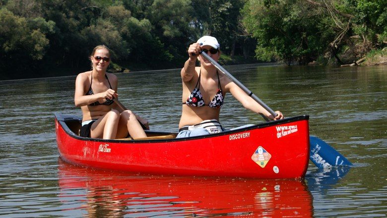 Zwei Frauen paddeln in einem roten Kanu auf einem Fluss, umgeben von grüner Natur.