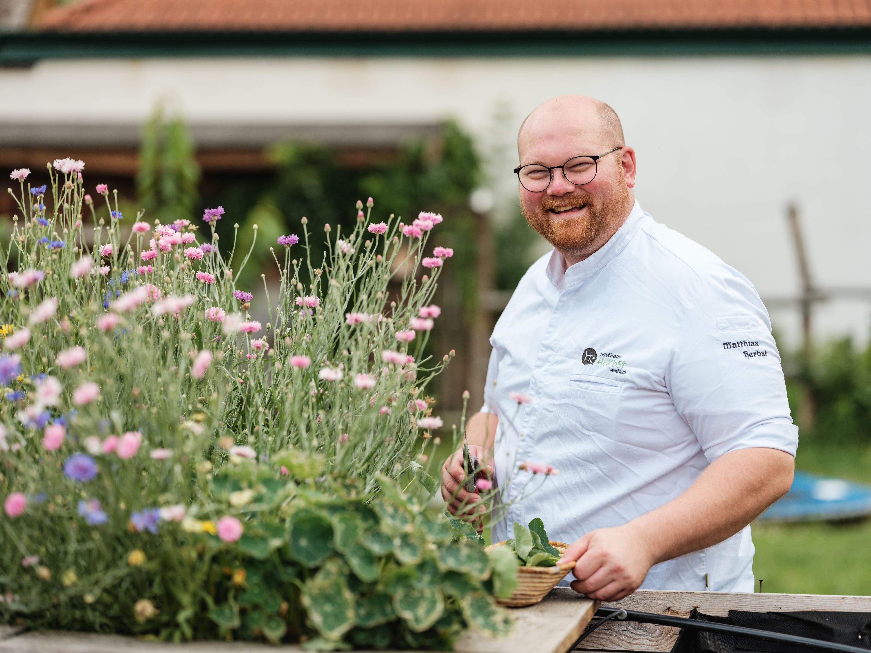 Ein lächelnder Mann in Kochkleidung steht neben blühenden Blumen im Freien.