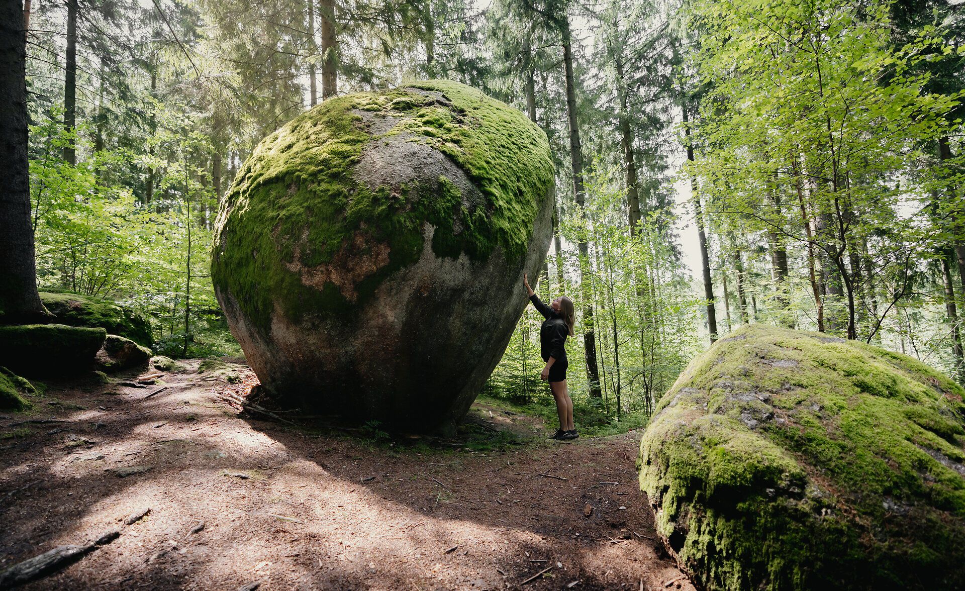 Person steht im Wald vor einem großen, kugelförmigen Felsen, der von Moos bedeckt ist; die sogenannte Weltkugel liegt in der Kraftarena Groß Gerungs, umgeben von hohen Bäumen.