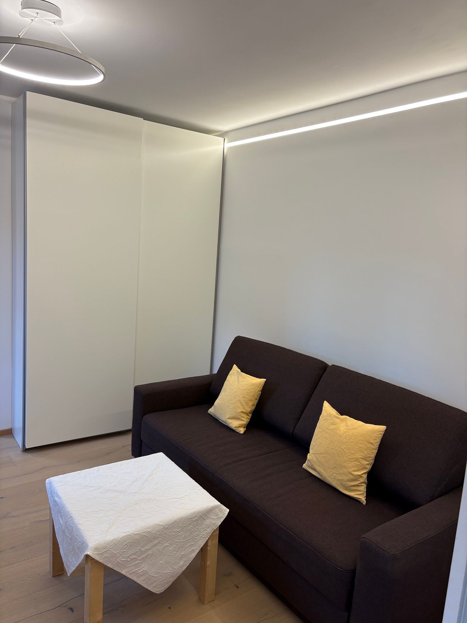 Modernes Wohnzimmer mit braunem Sofa, gelben Kissen und kleinem Tisch mit weißer Tischdecke.