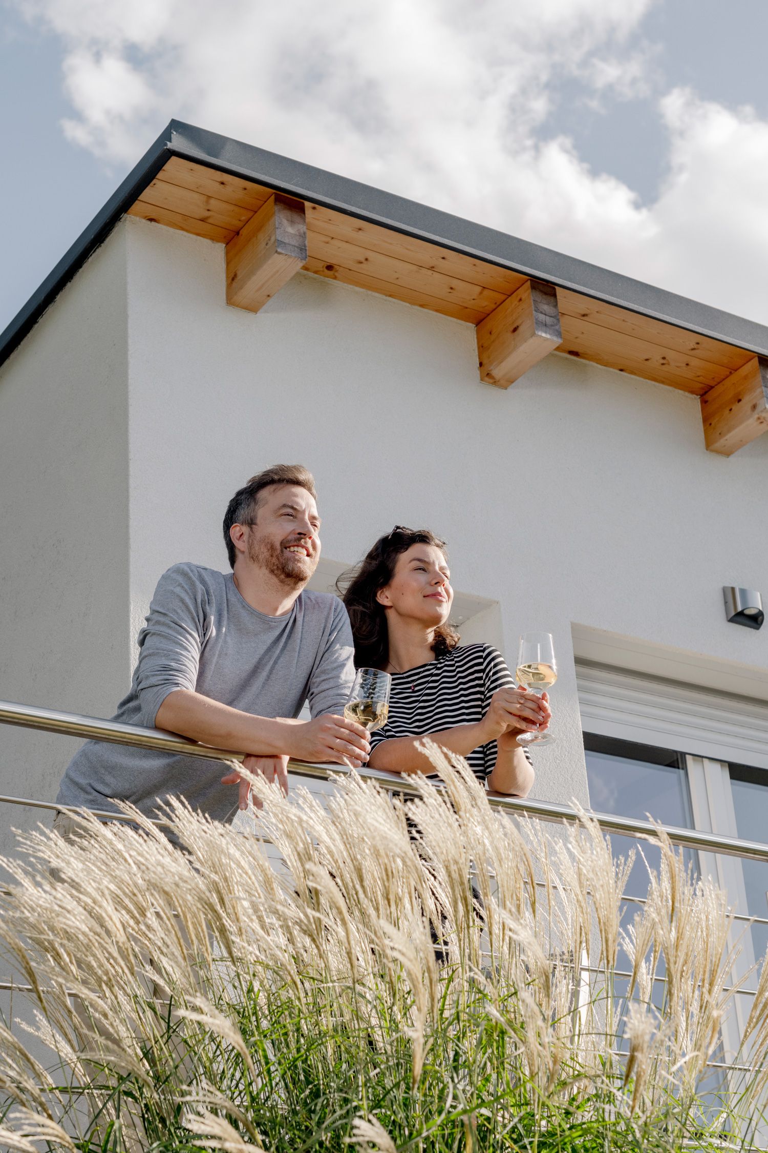 Ein Paar steht auf einem Balkon und hält Weingläser, mit einem modernen Gebäude im Hintergrund.
