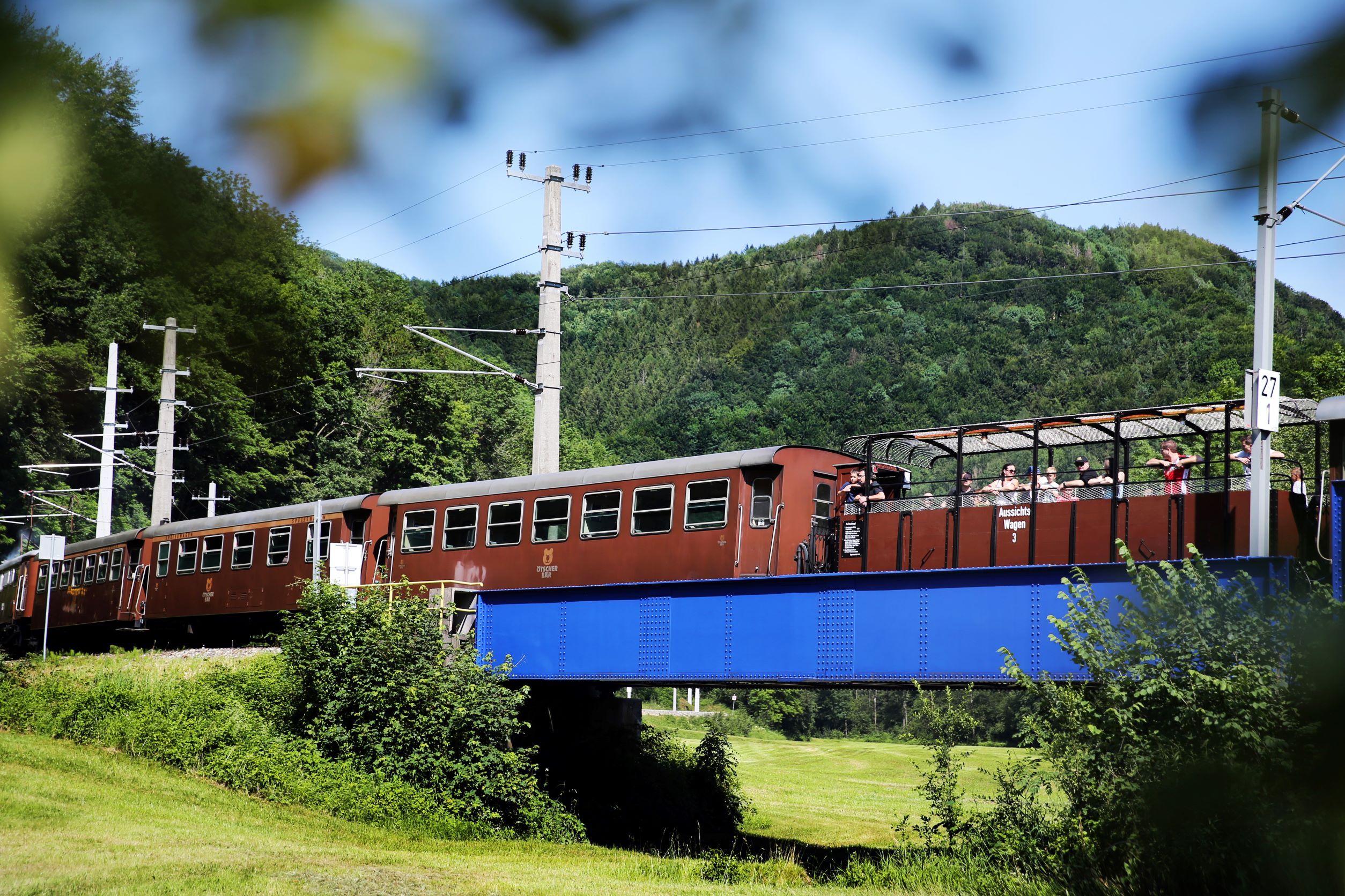 Ein Zug mit offenen Aussichtswagen fährt über eine blaue Brücke in einer grünen Landschaft.