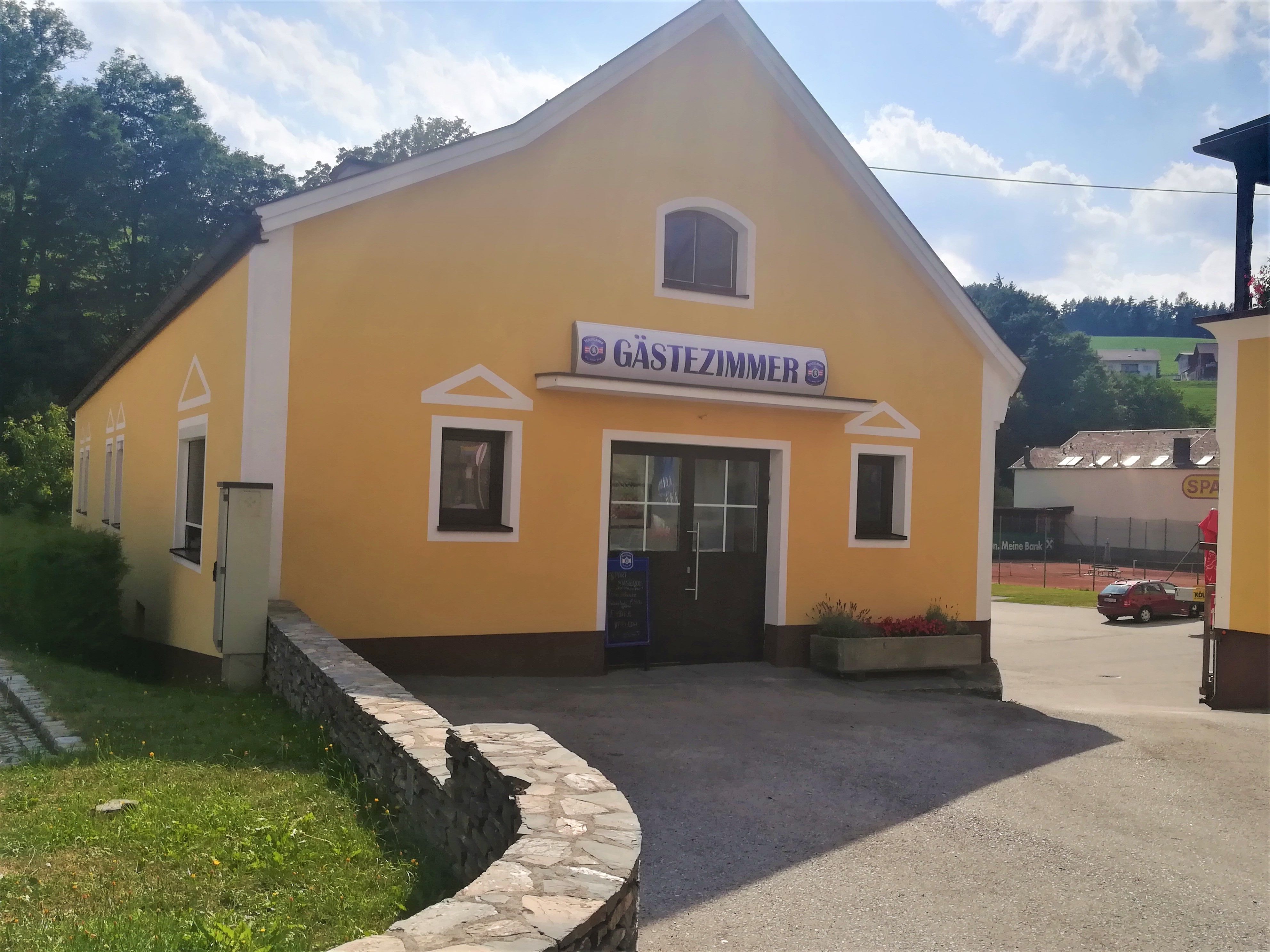 Gelbes Gebäude mit Schild 'Gästezimmer', umgeben von Bäumen und einem Parkplatz.