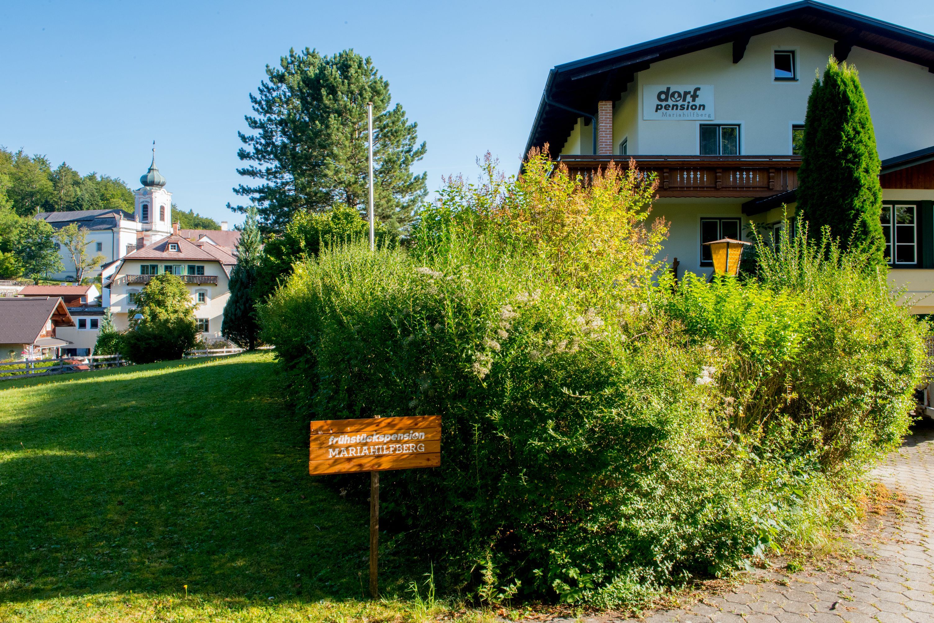 Ein Gebäude mit der Aufschrift 'Dorfpension' und einem Schild 'Frühstückspension Mariahilfberg' in einer grünen Umgebung.