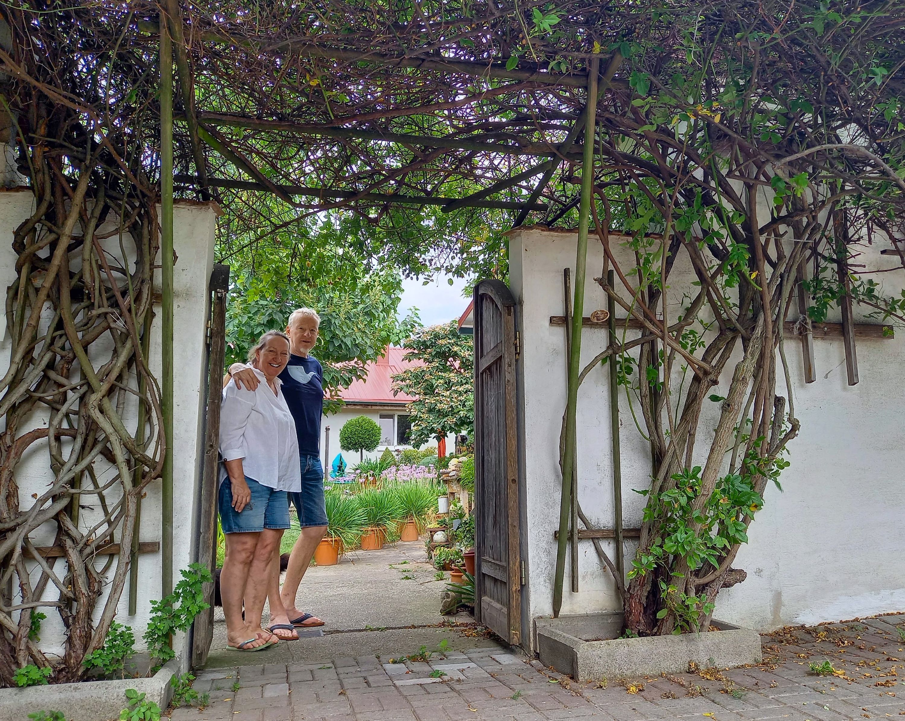 Zwei Personen stehen lächelnd unter einem mit Pflanzen bewachsenen Torbogen in einem Garten.