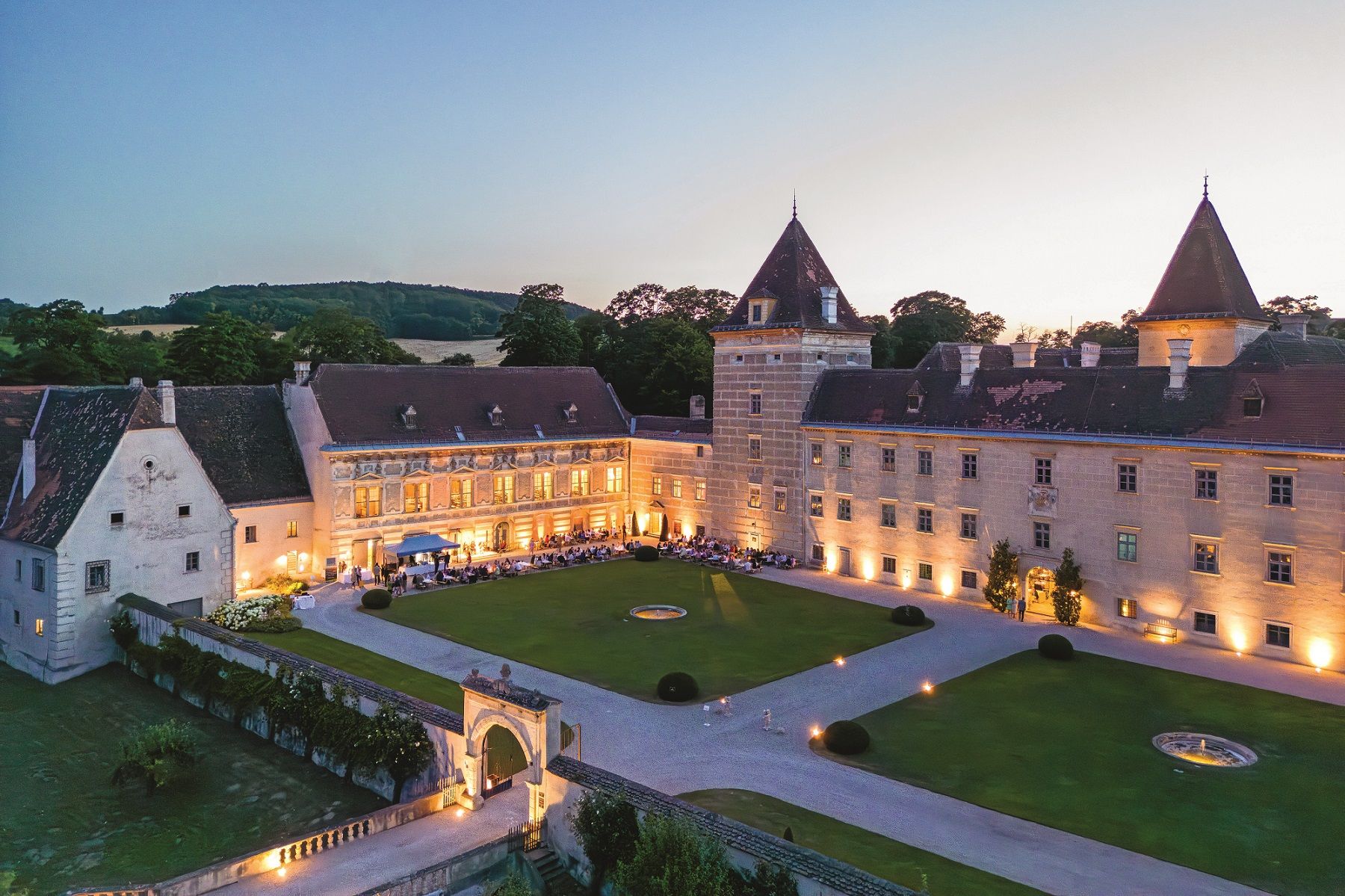 Luftaufnahme von Schloss Walpersdorf bei Dämmerung, beleuchtet mit Gästen bei einem Gartenfest im Innenhof.