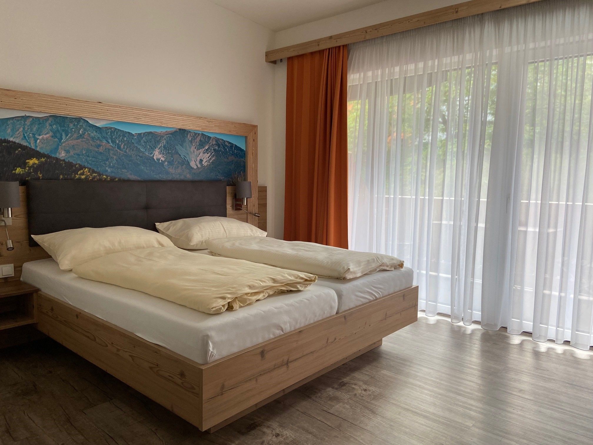 Modernes Doppelzimmer mit großem Bett, Bergbild an der Wand und großen Fenstern mit Vorhängen.