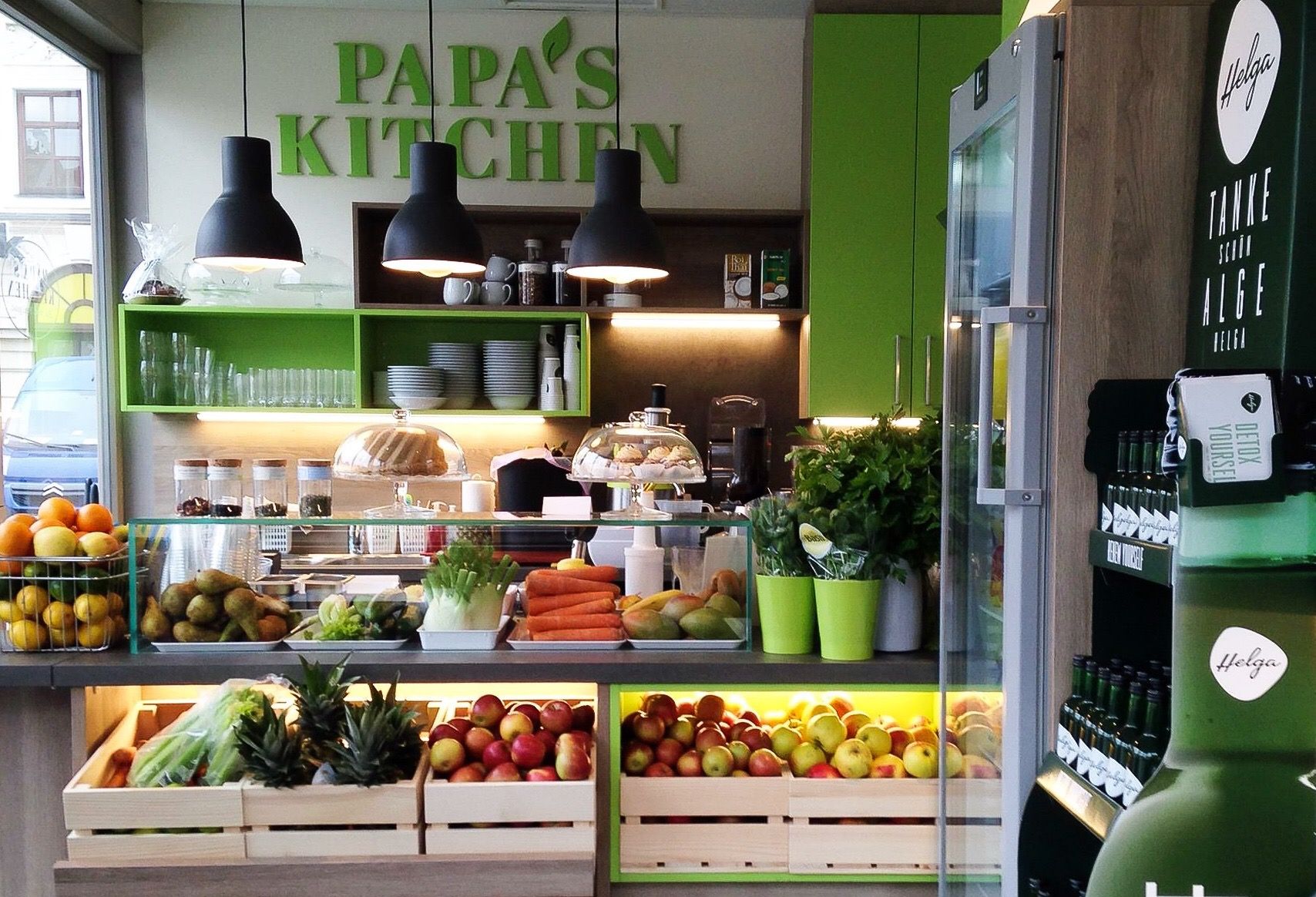 Innenansicht eines modernen Lokals mit frischem Obst und Gemüse, grüner Dekoration und dem Schriftzug 'Papa's Kitchen'.