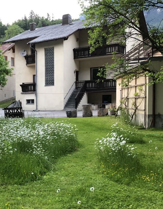 Ein zweistöckiges Haus mit Balkon und Treppe, umgeben von einem grünen Garten mit Blumen.