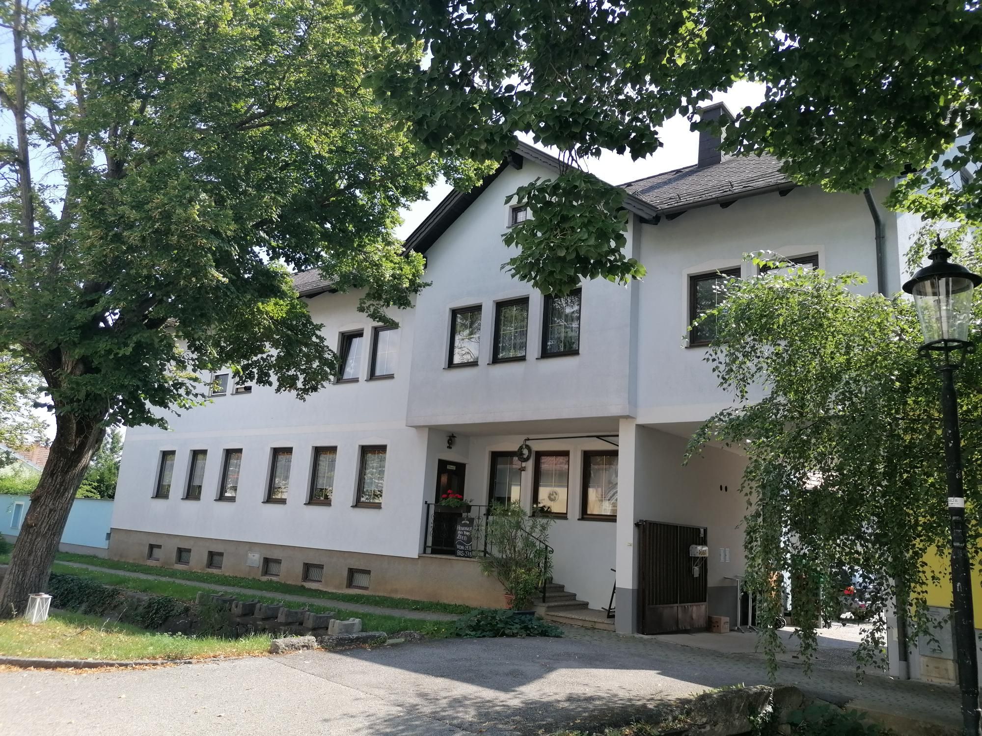 Außenansicht eines weißen Gebäudes mit vielen Fenstern, umgeben von Bäumen.