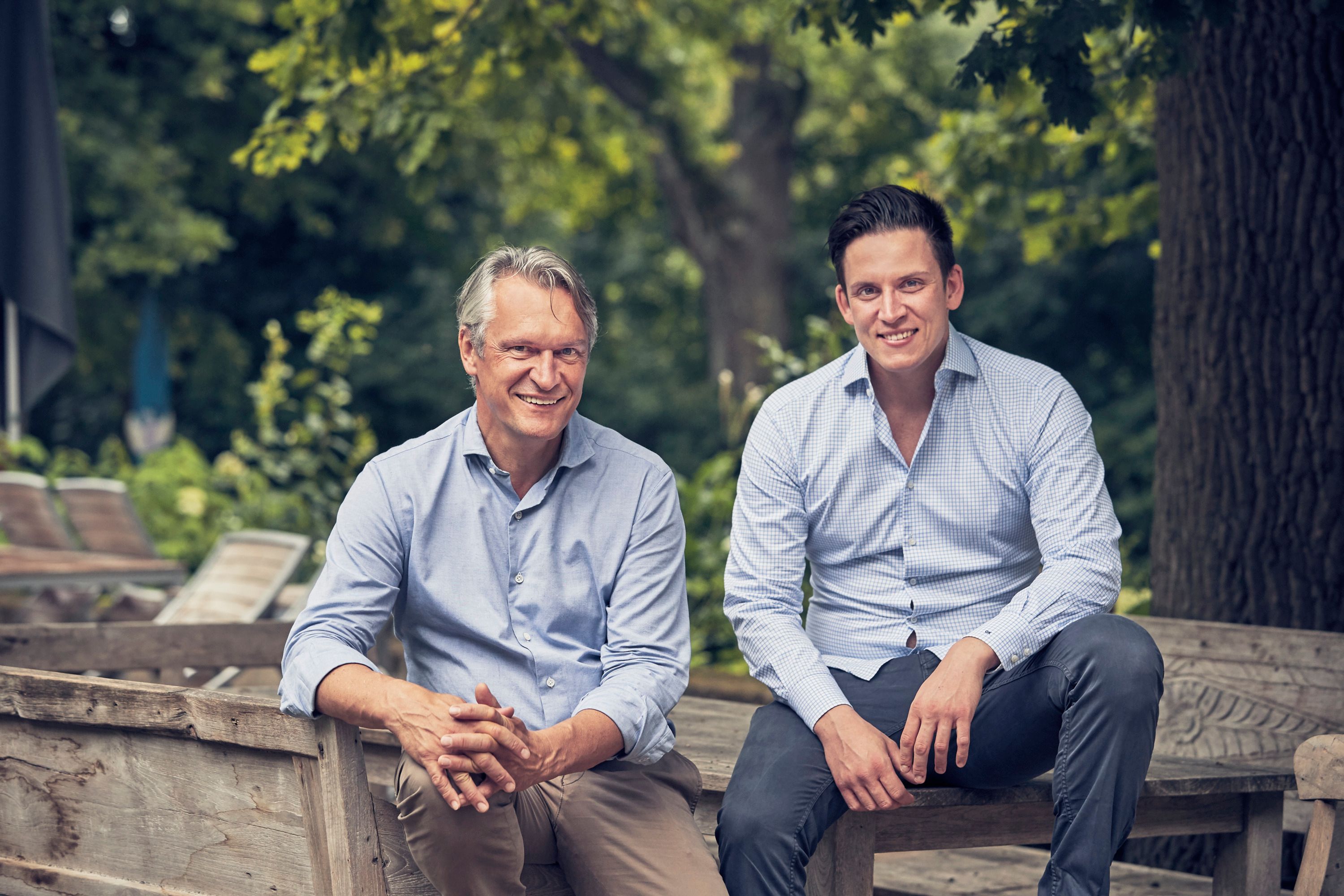 Zwei Männer sitzen auf einer Holzbank im Freien, umgeben von Bäumen.