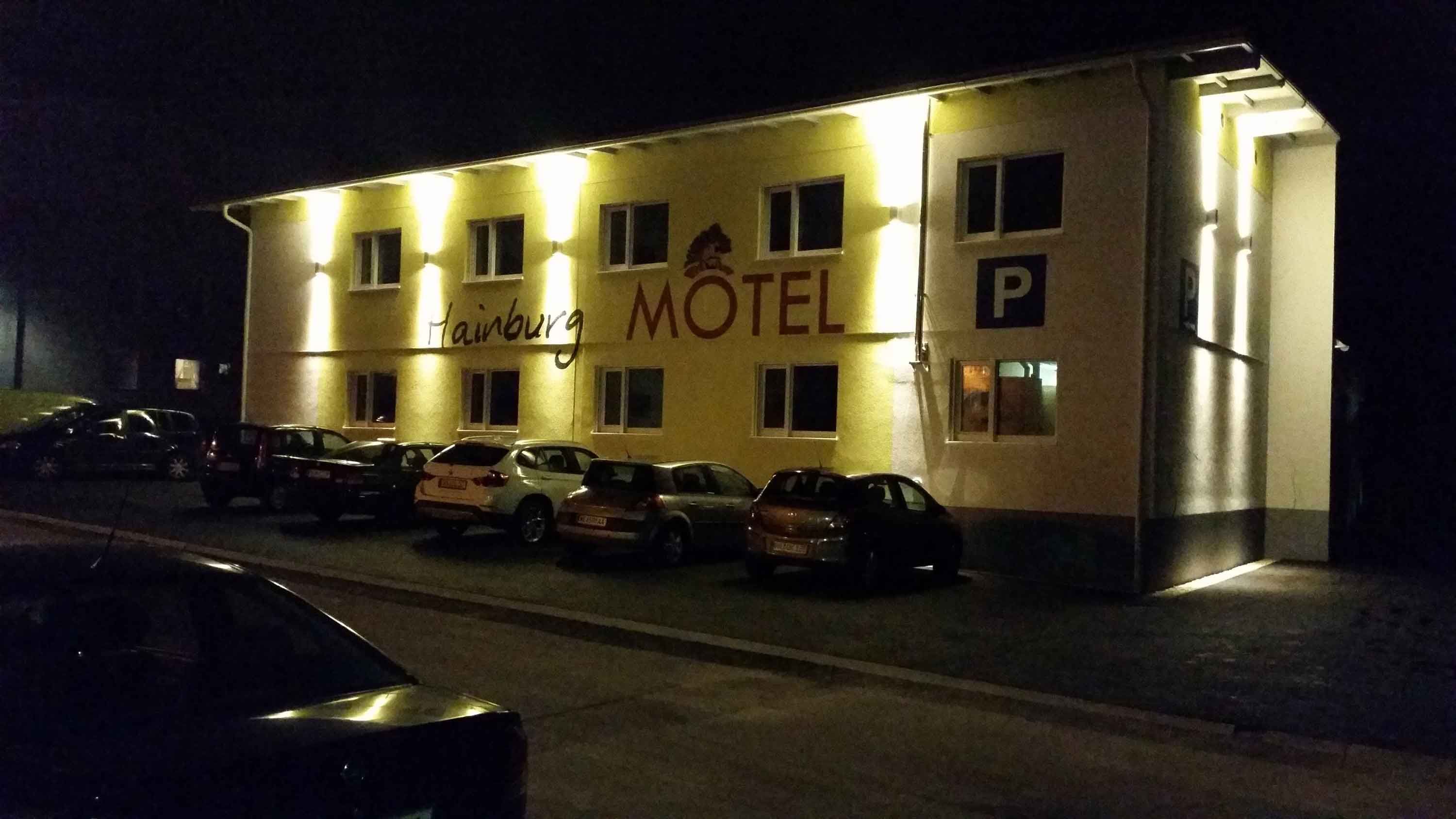 Nachtaufnahme eines beleuchteten Motels mit dem Schriftzug 'Hainburg Motel' und geparkten Autos davor.