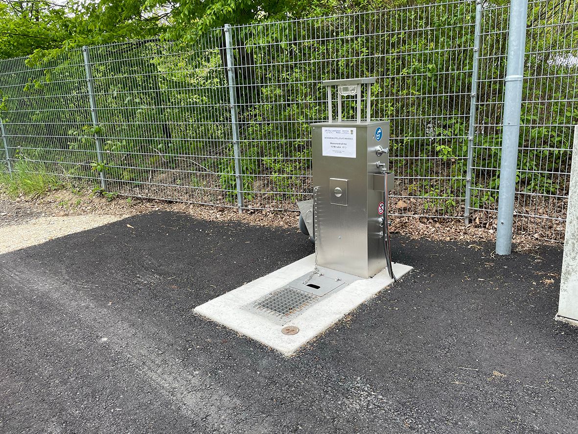Metallene Wasserversorgungsstation neben einem Zaun auf Asphalt.