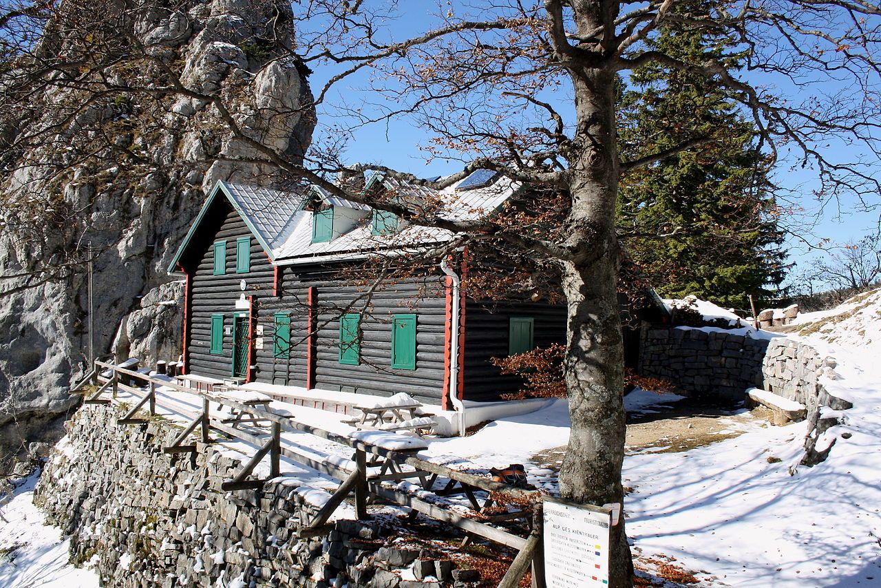 Eine Berghütte aus Holz mit grünen Fensterläden, umgeben von Schnee und Felsen, unter einem klaren blauen Himmel.