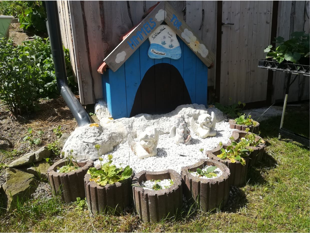 Kleines blaues Häuschen mit Engelfiguren und Pflanzen in einem Garten.