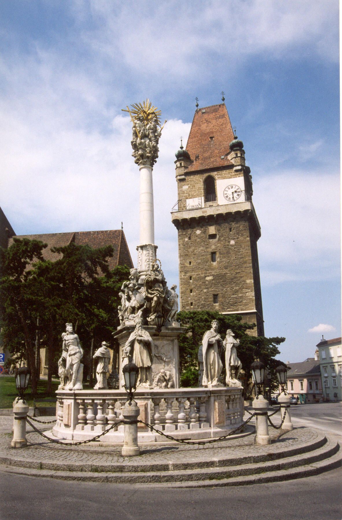 Historischer Wehrturm mit Uhr und barocker Säule im Vordergrund.