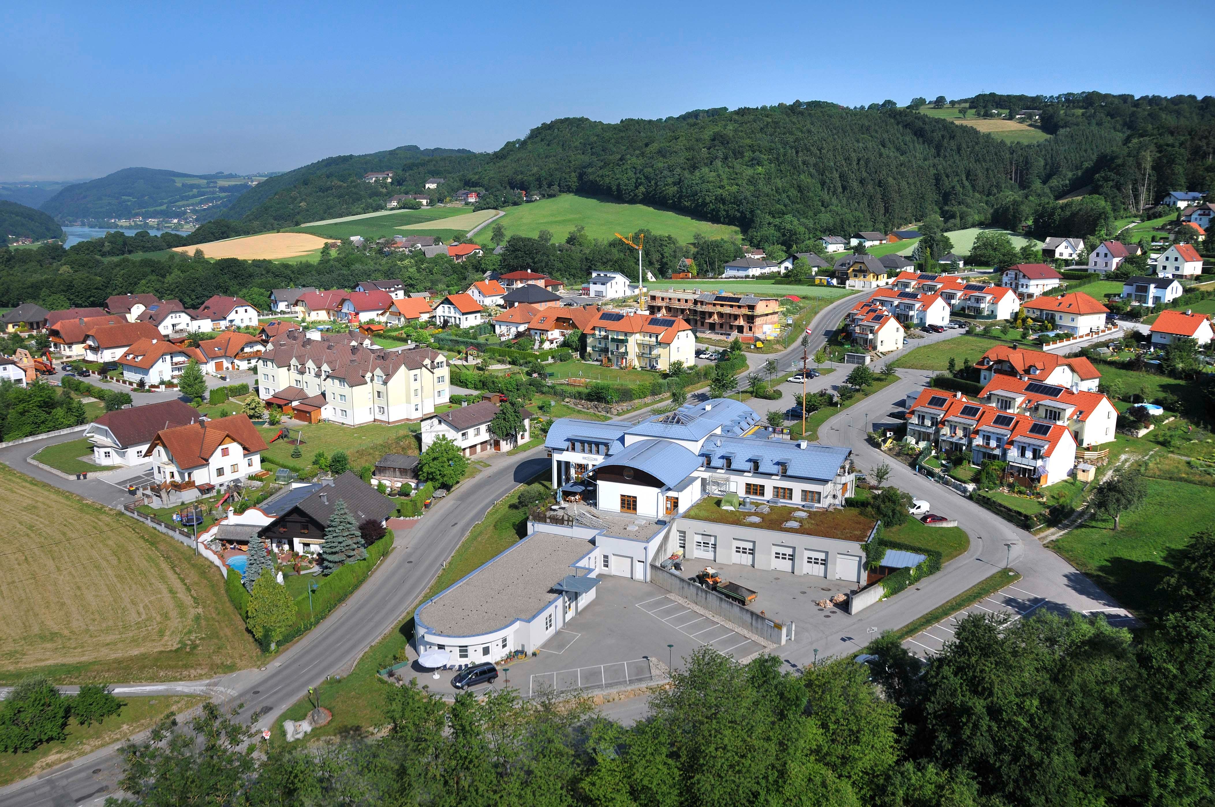 Luftaufnahme eines Dorfes mit Gemeindezentrum und umliegenden Häusern in einer grünen Hügellandschaft.