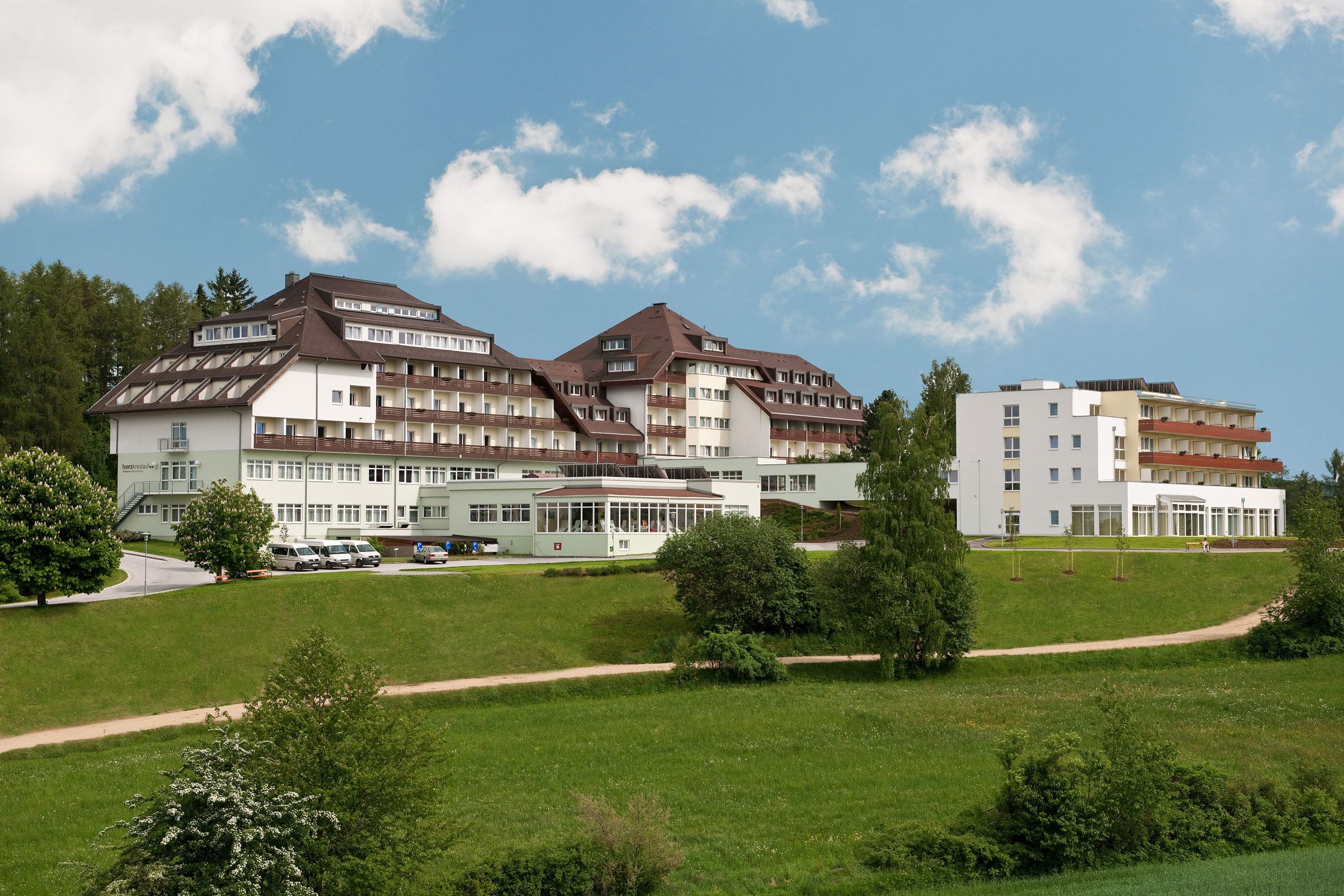 Großes Gebäude mit mehreren Stockwerken und Balkonen, umgeben von grüner Landschaft und Bäumen, unter blauem Himmel.