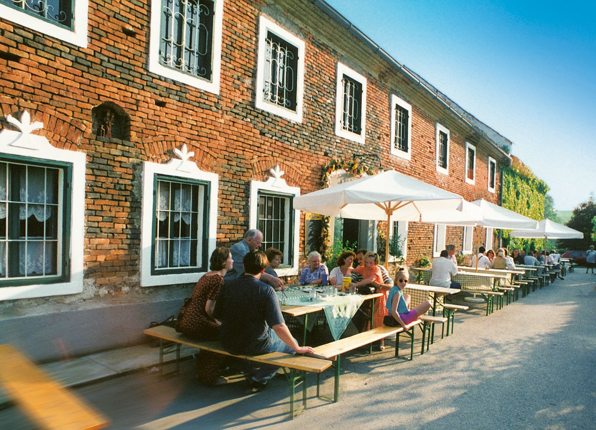 Menschen sitzen an Tischen vor einem Ziegelgebäude mit Sonnenschirmen.