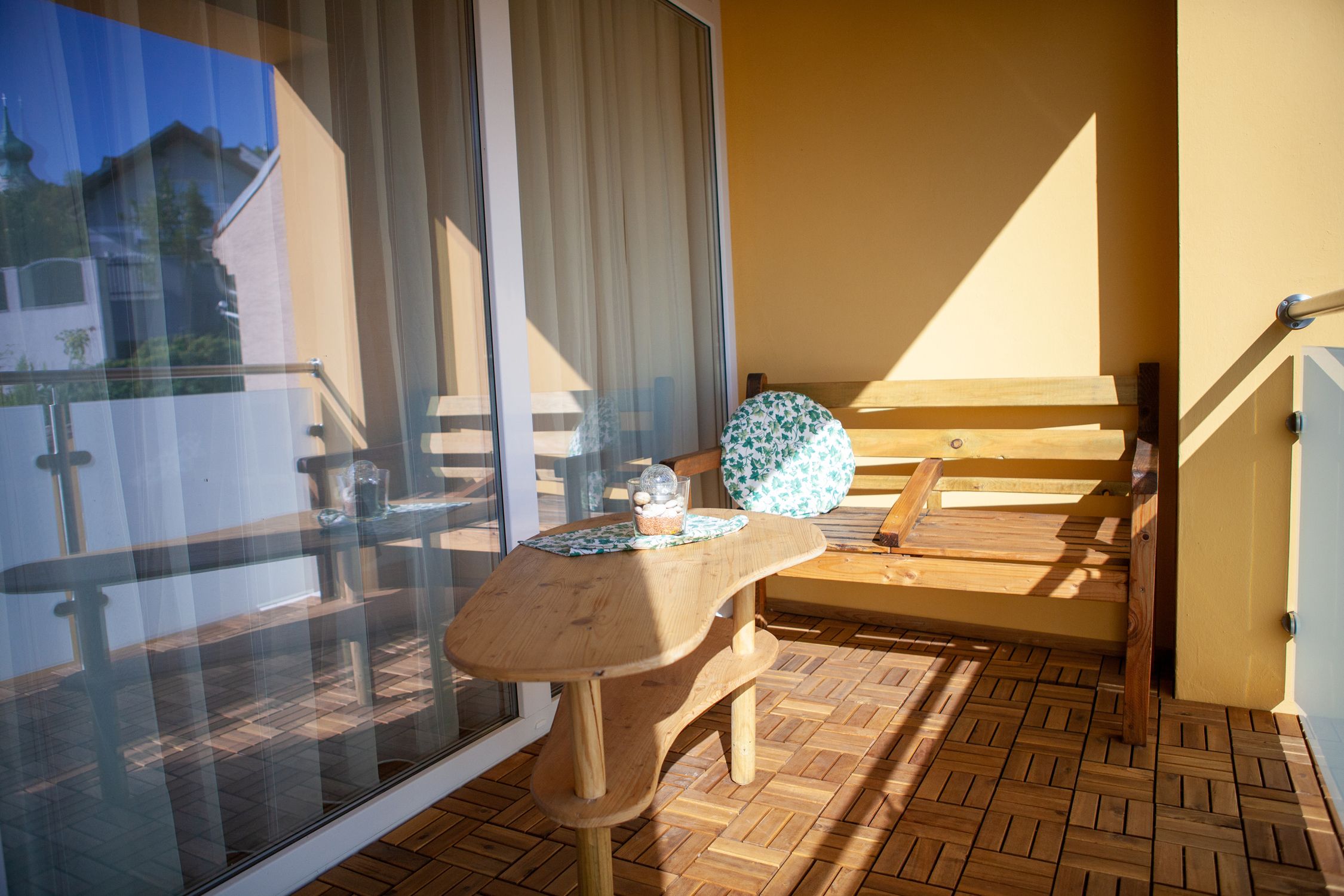 Ein Balkon mit Holzboden, einem Holztisch und einer Holzbank mit einem runden Kissen. Im Hintergrund ist ein Glasgeländer und ein Gebäude zu sehen.