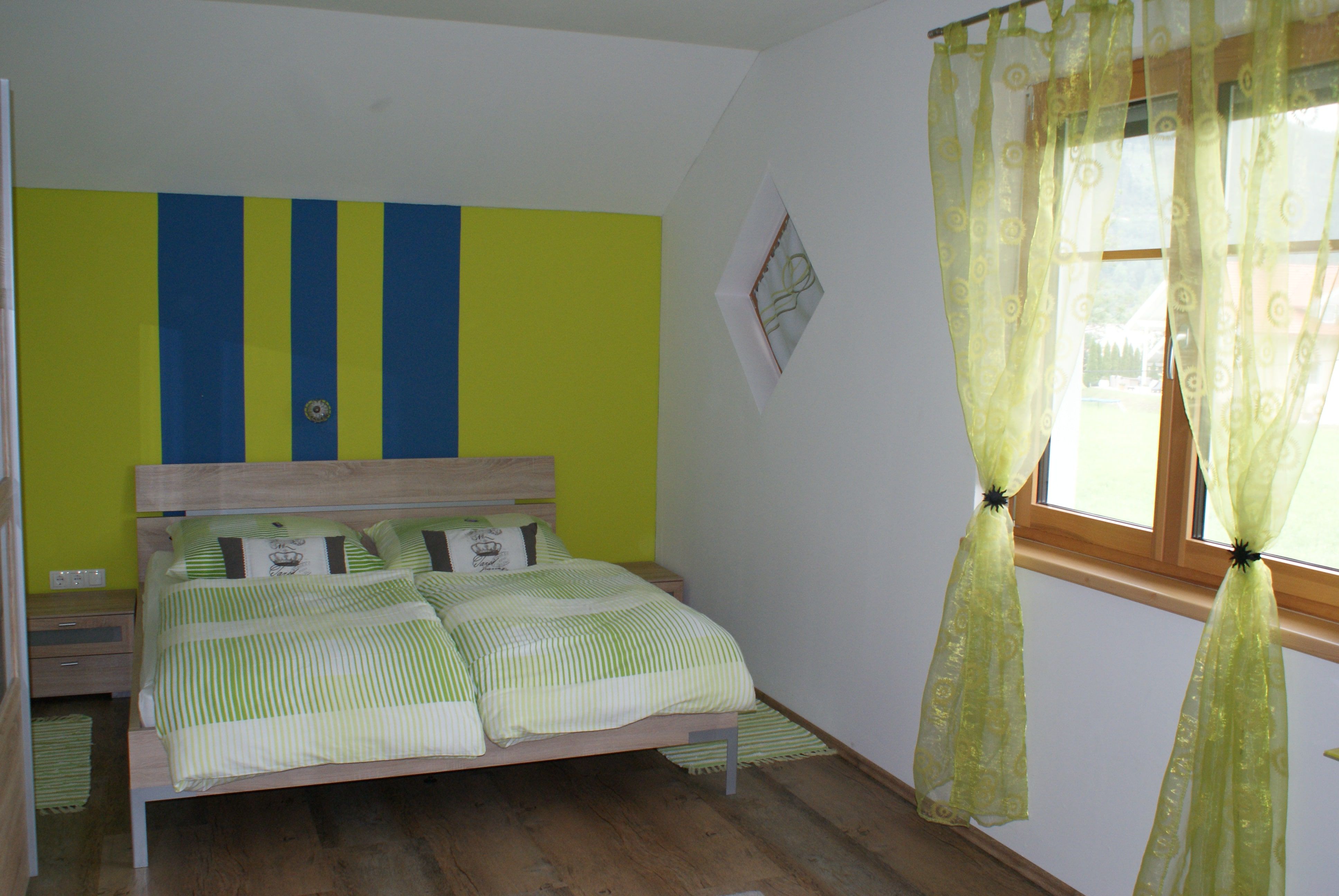 Schlafzimmer mit Doppelbett, grün-gelb gestreifte Wand, grüne Vorhänge, Holzboden.