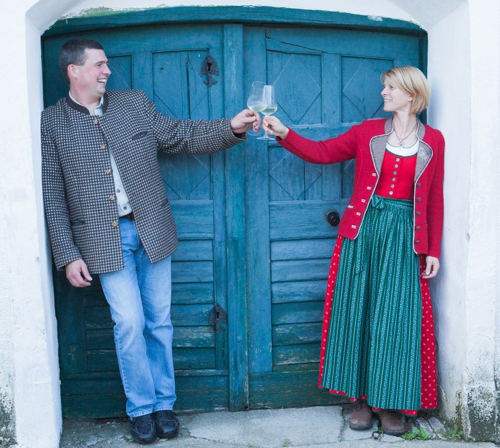 Ein Mann und eine Frau stoßen mit Weingläsern an, vor einer blauen Holztür.
