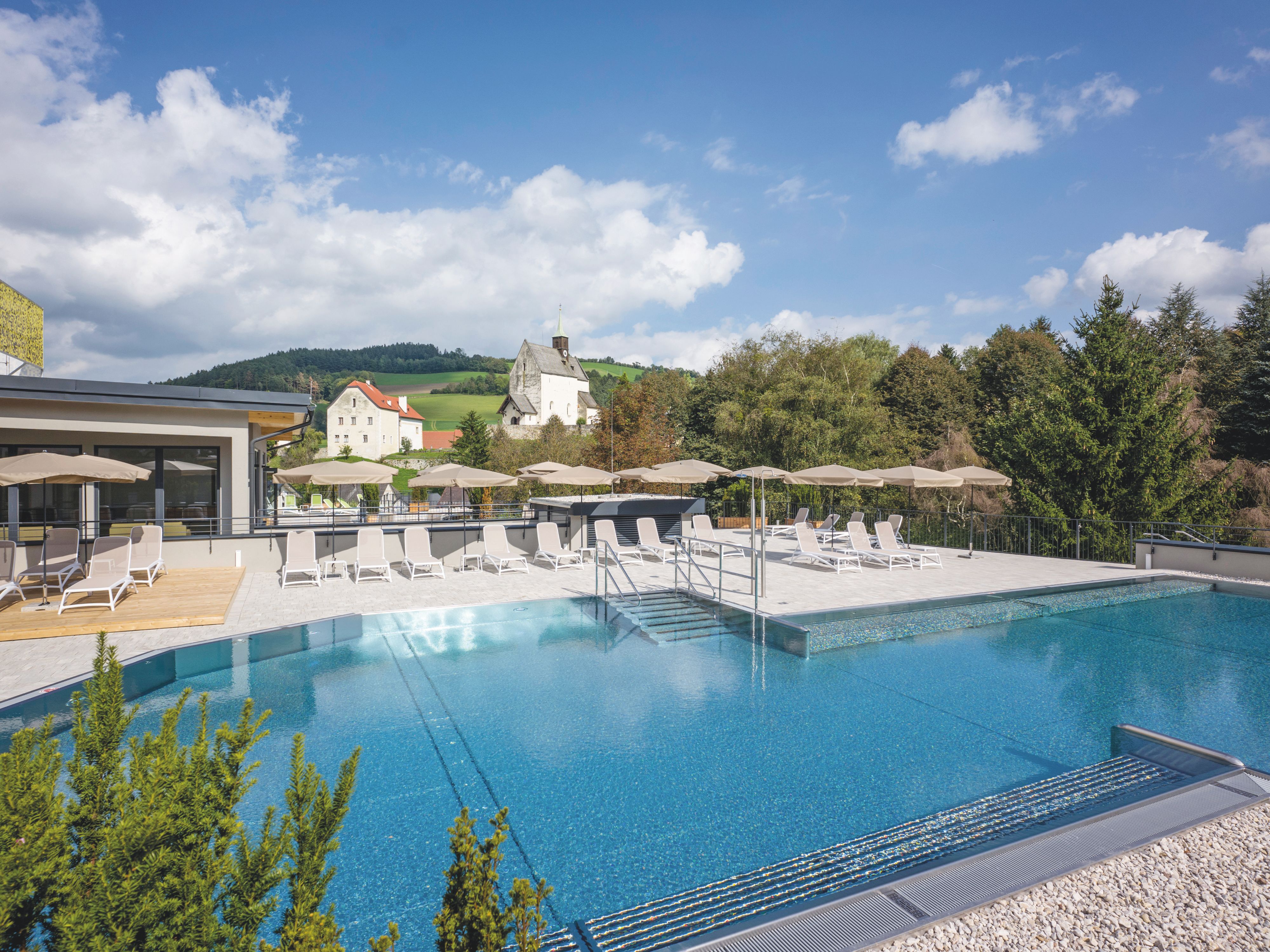 Außenpool mit Liegestühlen und Sonnenschirmen, im Hintergrund eine Kirche und grüne Hügel.