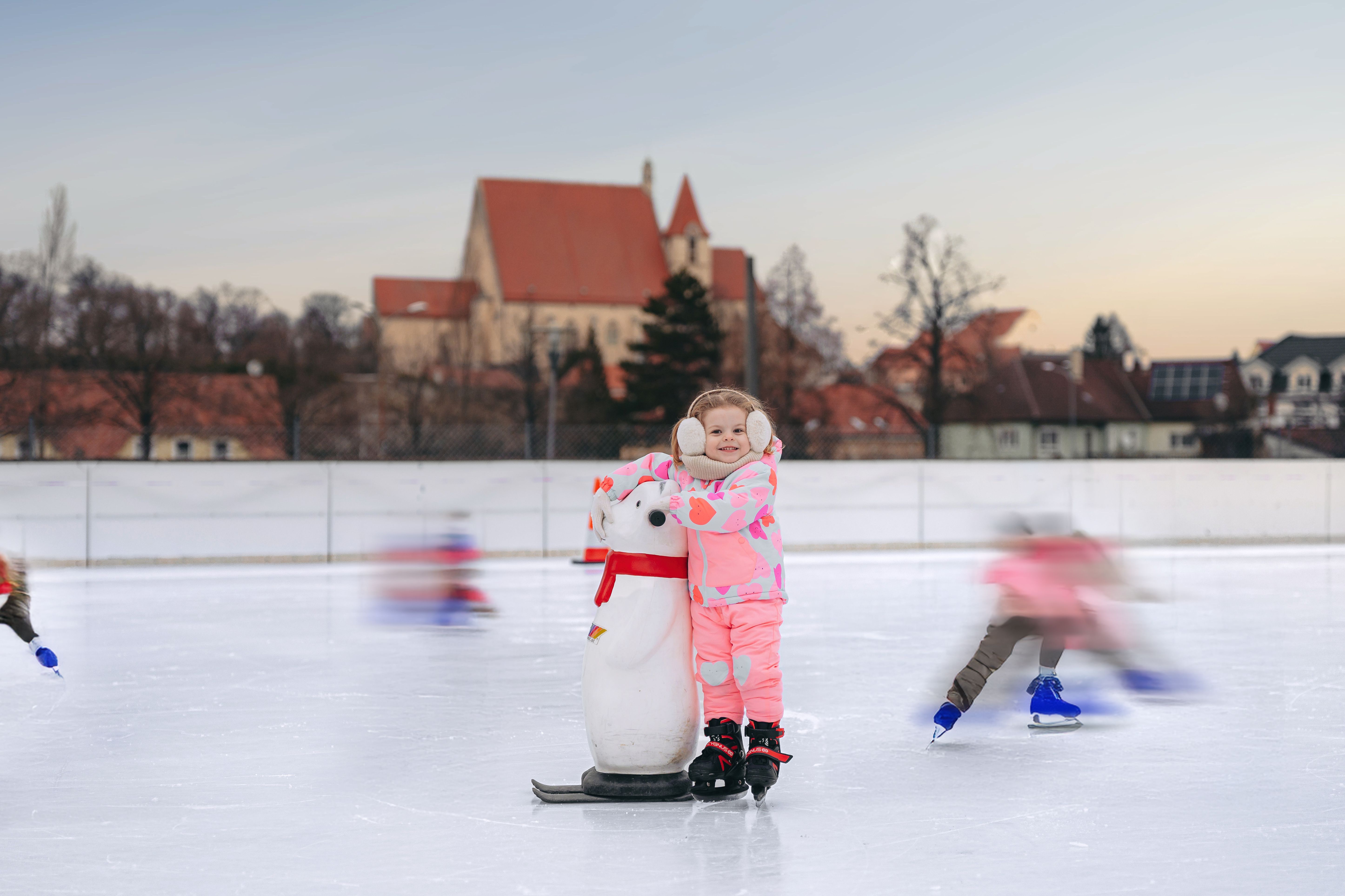 Ein Kind in pinker Kleidung steht auf einer Eisbahn mit einem Pinguin-Hilfsmittel. Im Hintergrund ist eine Kirche zu sehen.