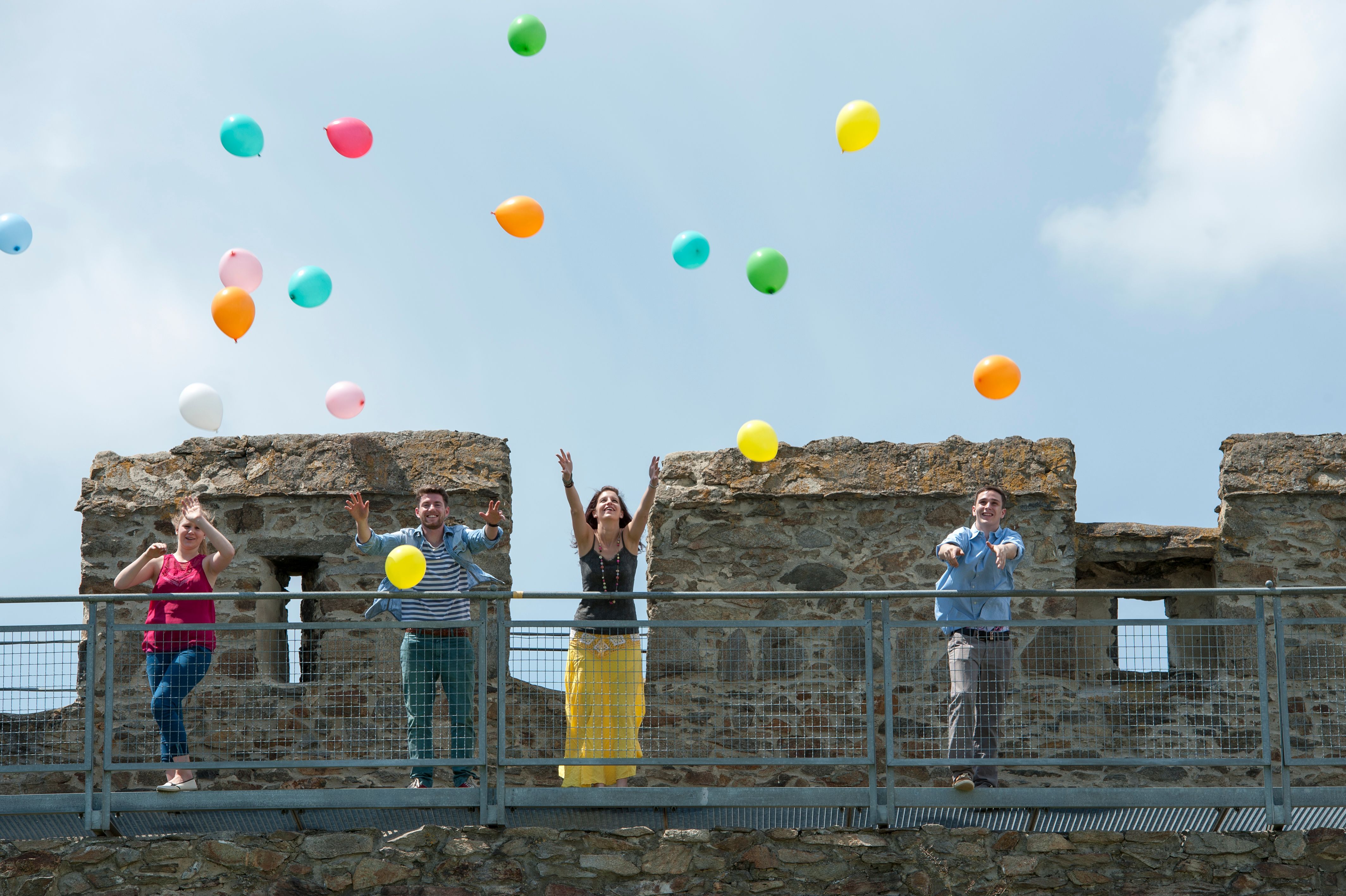 Vier Personen werfen bunte Luftballons von einer Burgmauer in die Luft.