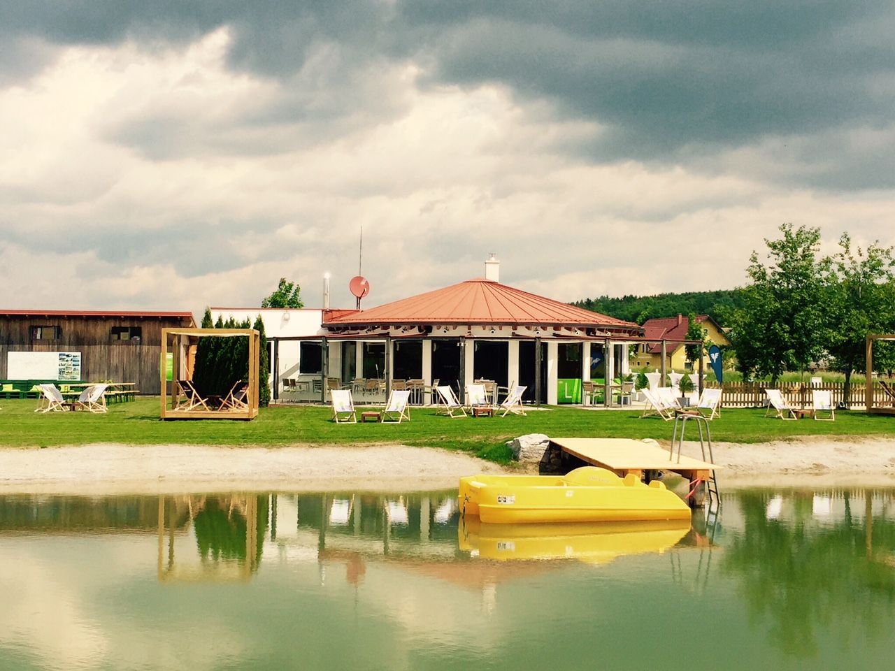 Ein Erholungsteich mit gelbem Tretboot, Liegestühlen und einem Pavillon im Hintergrund.